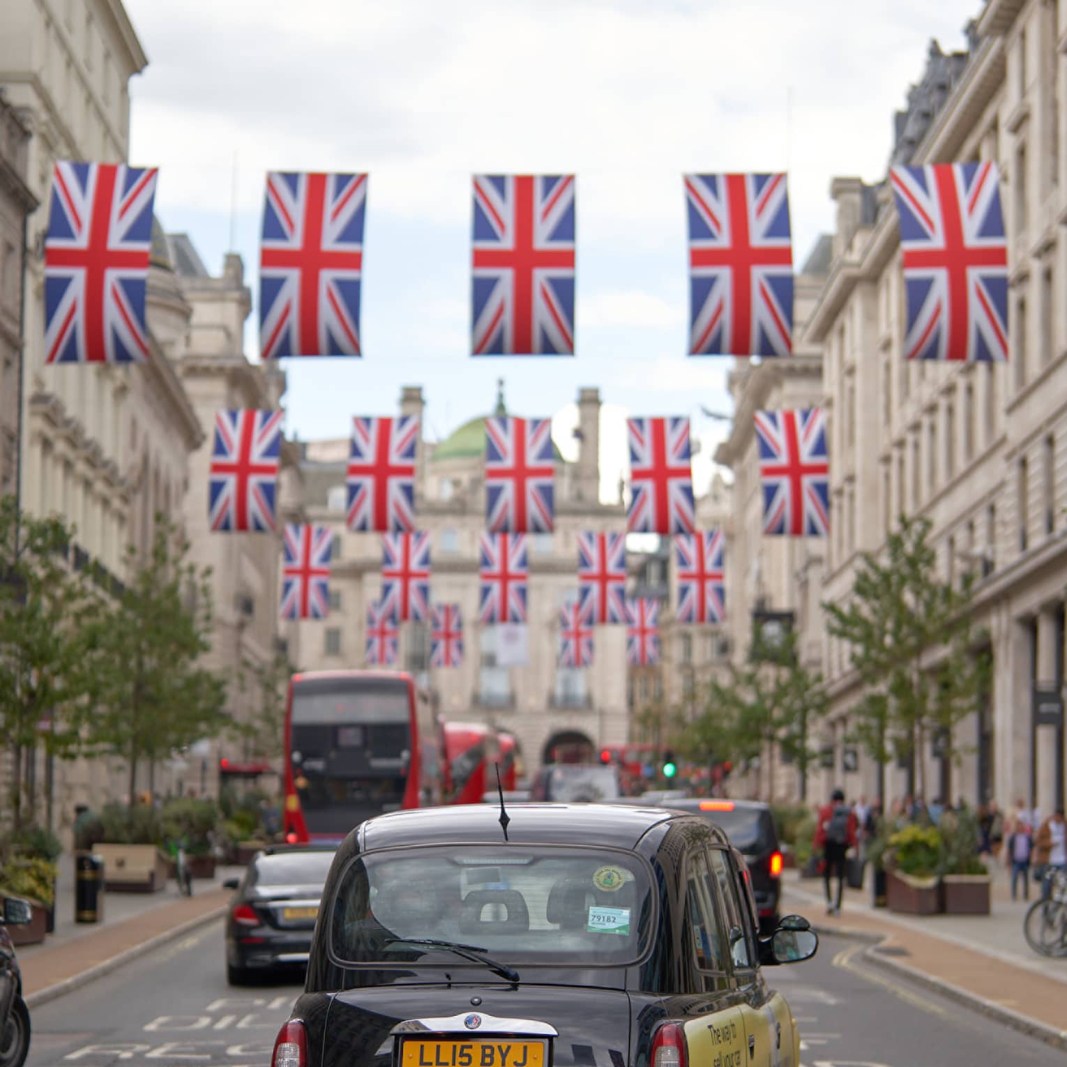 Rear view of London-style black cab, double-decker buses and other cars on road strung across with rows of Union Jack flags.