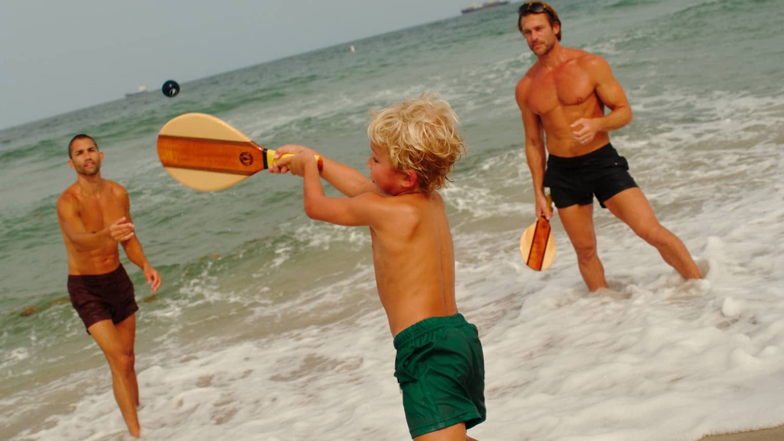 A young shild plays paddle ball in the surf with two adults