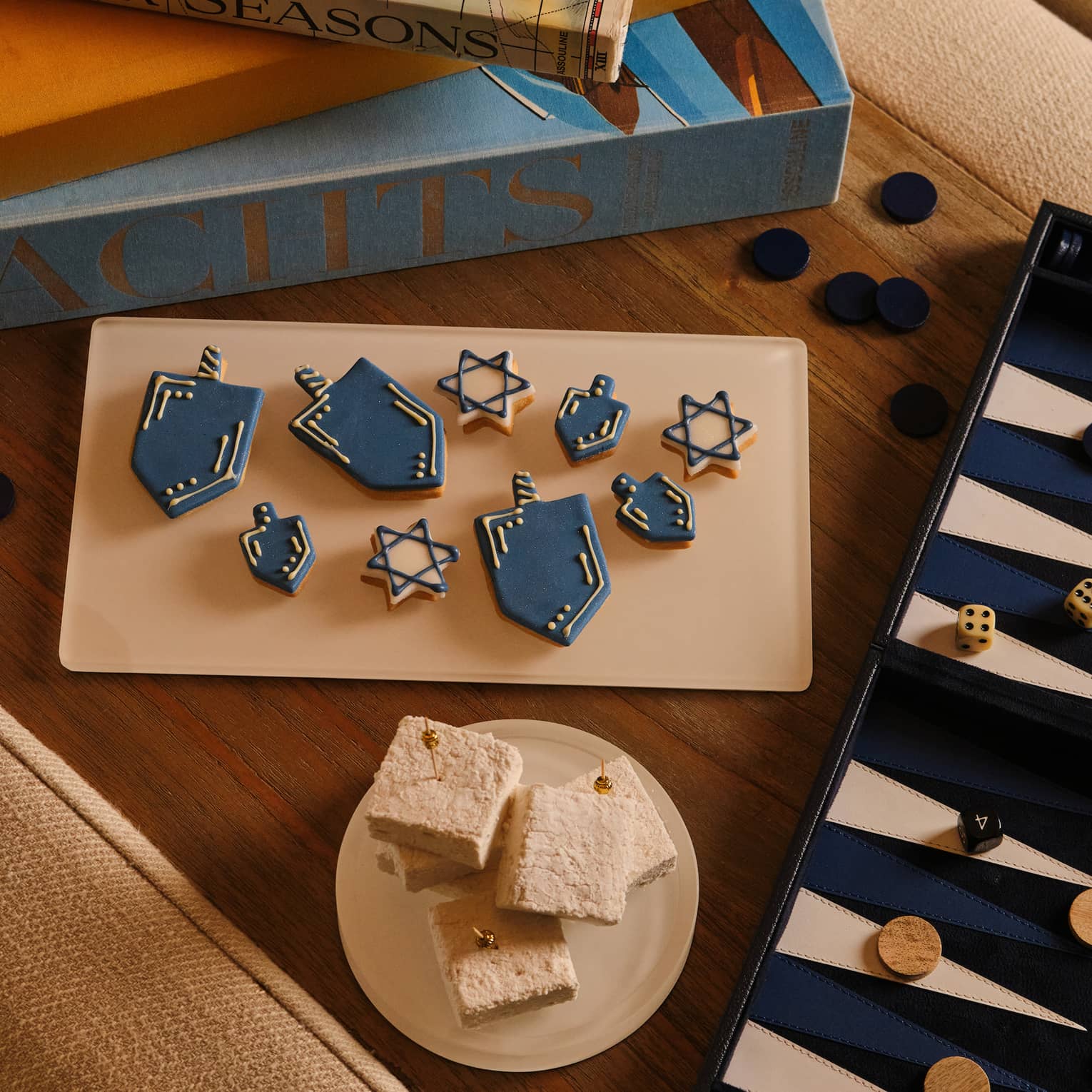 Small desserts shapped like dreidels and Stars of David set next to a blue and white backgammon board and astack of books