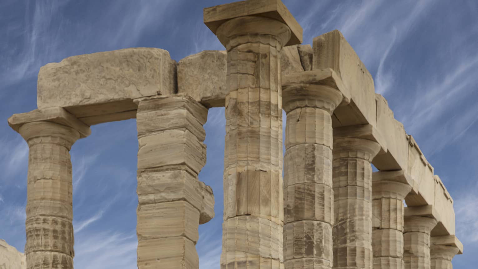 Side view of ancient ruins with formidable Doric columns on a stone foundation, cloud-streaked sky and hilly islands beyond.