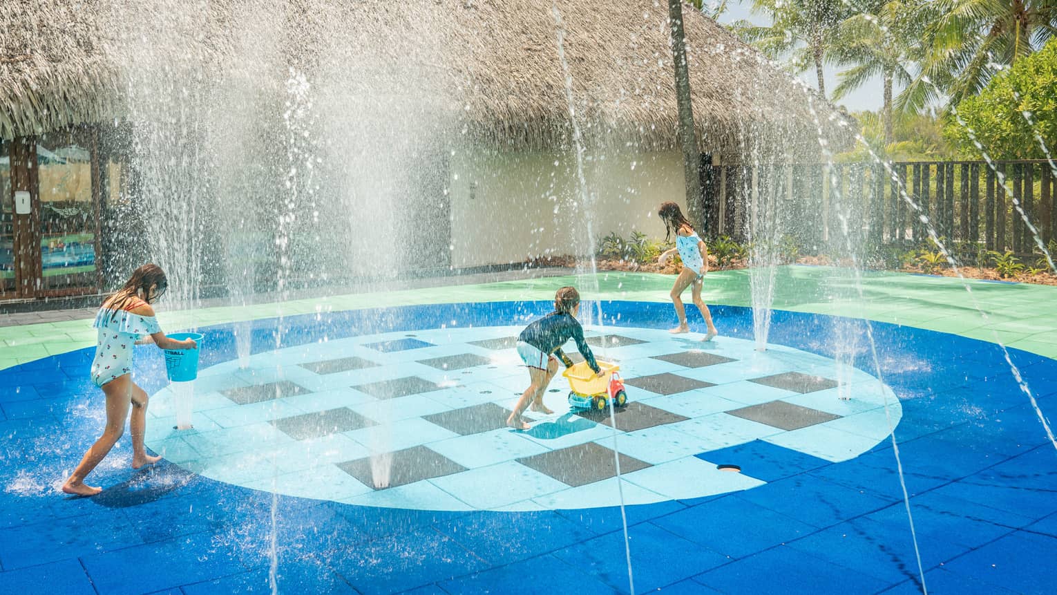 Group of children in swimsuits play on splash pad next to grass-roof building