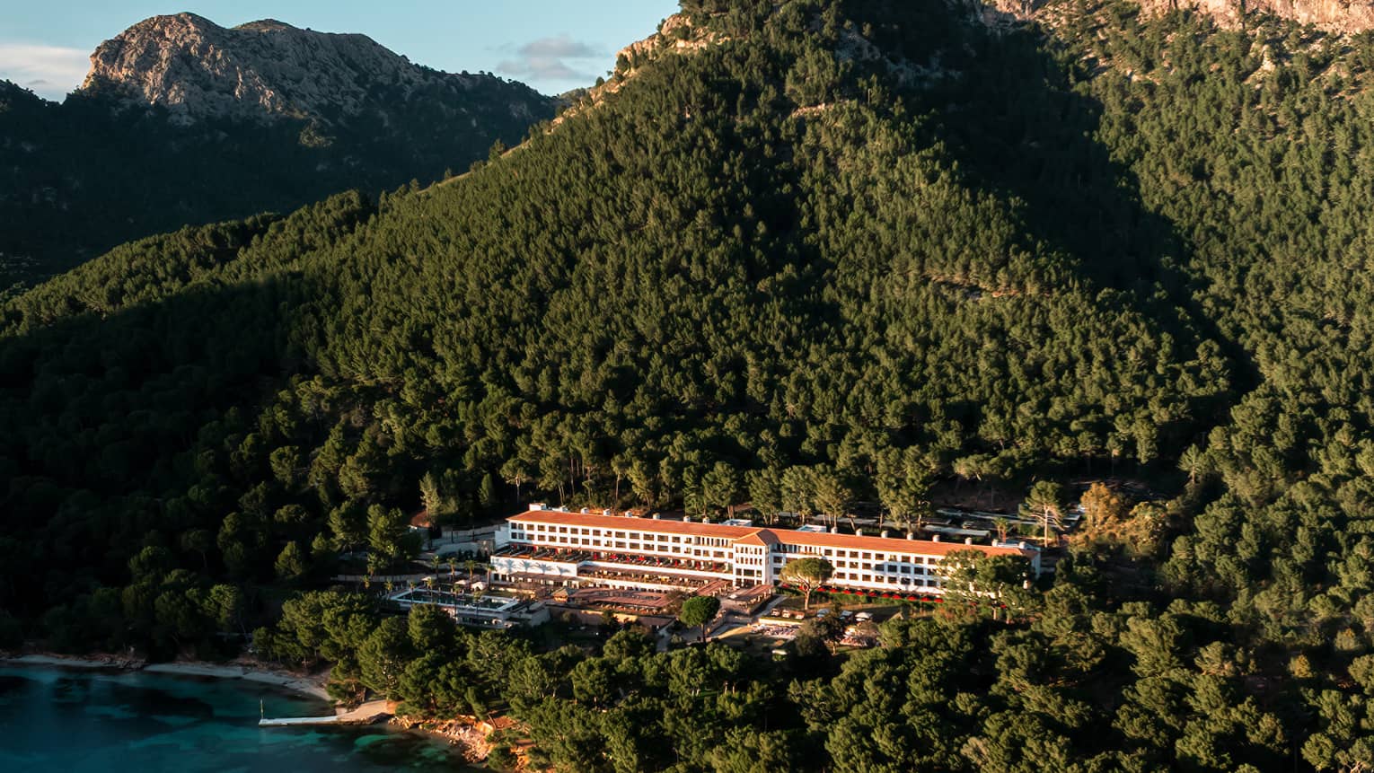 An aerial view of a wooded, mountainous coastline with a resort building among the trees, blue water in the foreground, and mountain views and blue sky in the background