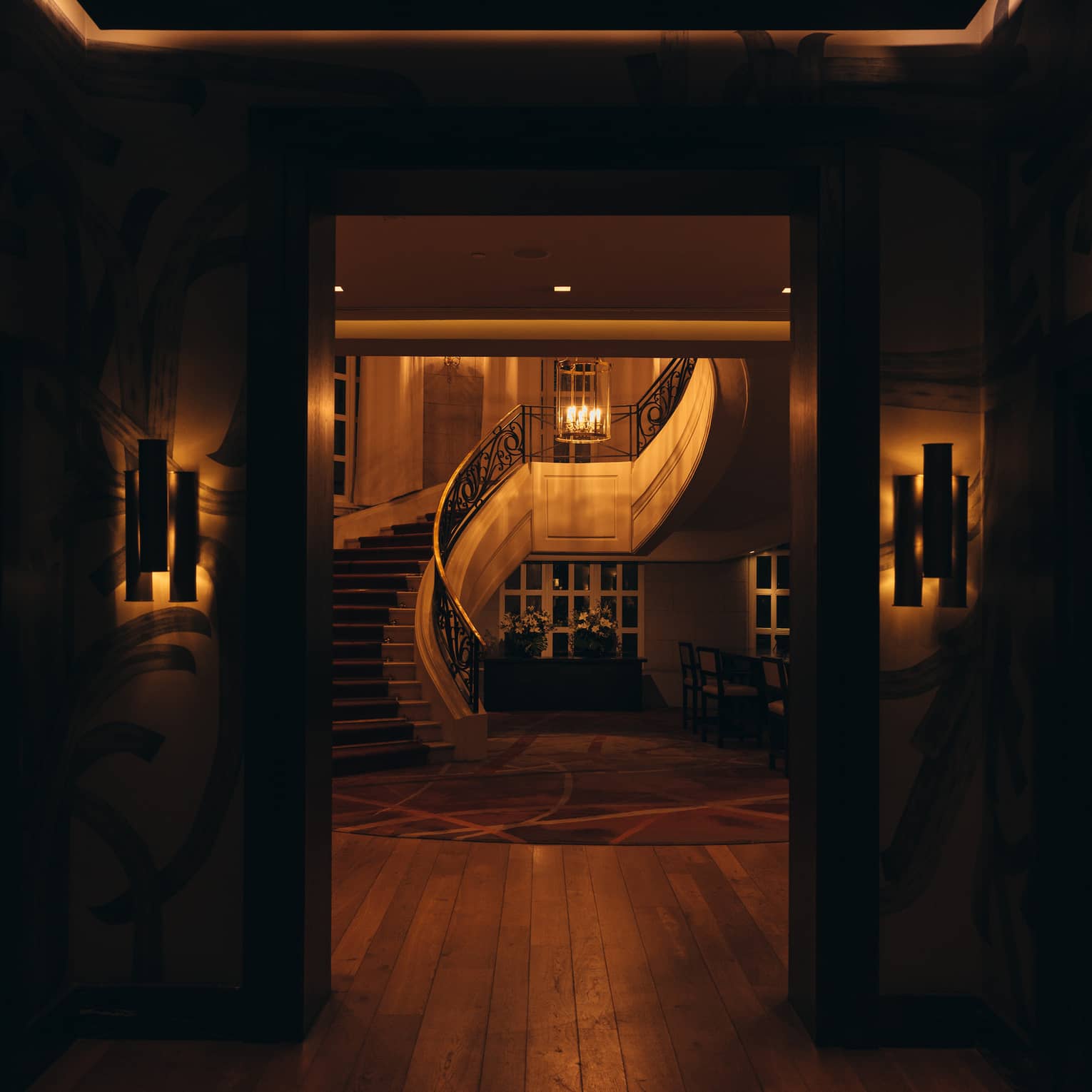 An interior space in the warm, low light of evening, featuring wall sconce lighting and a curved staircase and chandelier seen through a doorway.