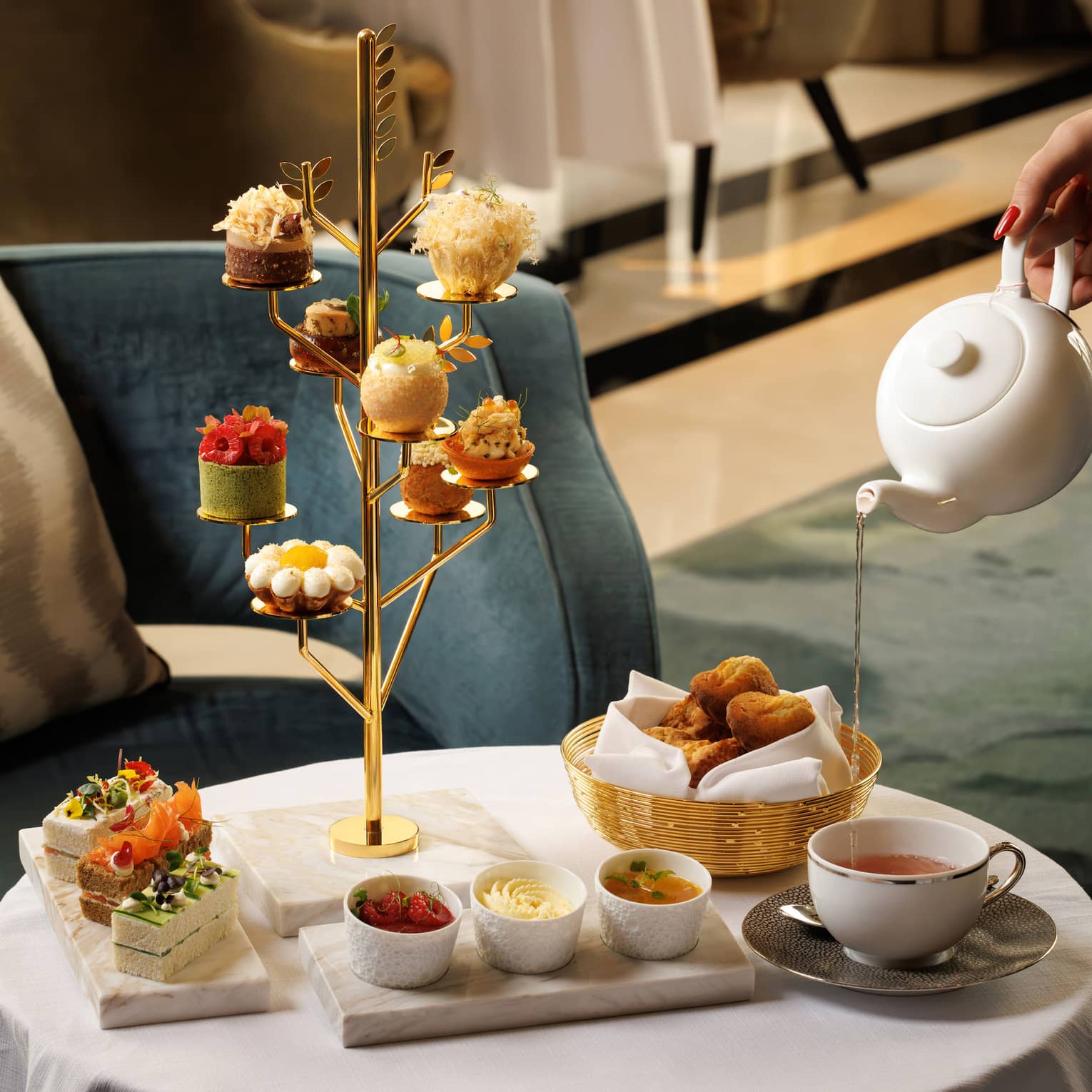 Golden tree-shaped stand holds a small pastry on each branch, set next to a marble plate of finger sandwiches, basket of bread, three small white bowls and a cup of tea