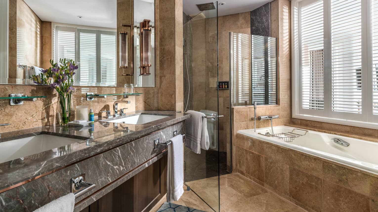 A marble bathroom with double sinks, large vanity, glass-enclosed shower and soaking tub by the window. Fresh flowers add a touch of elegance.