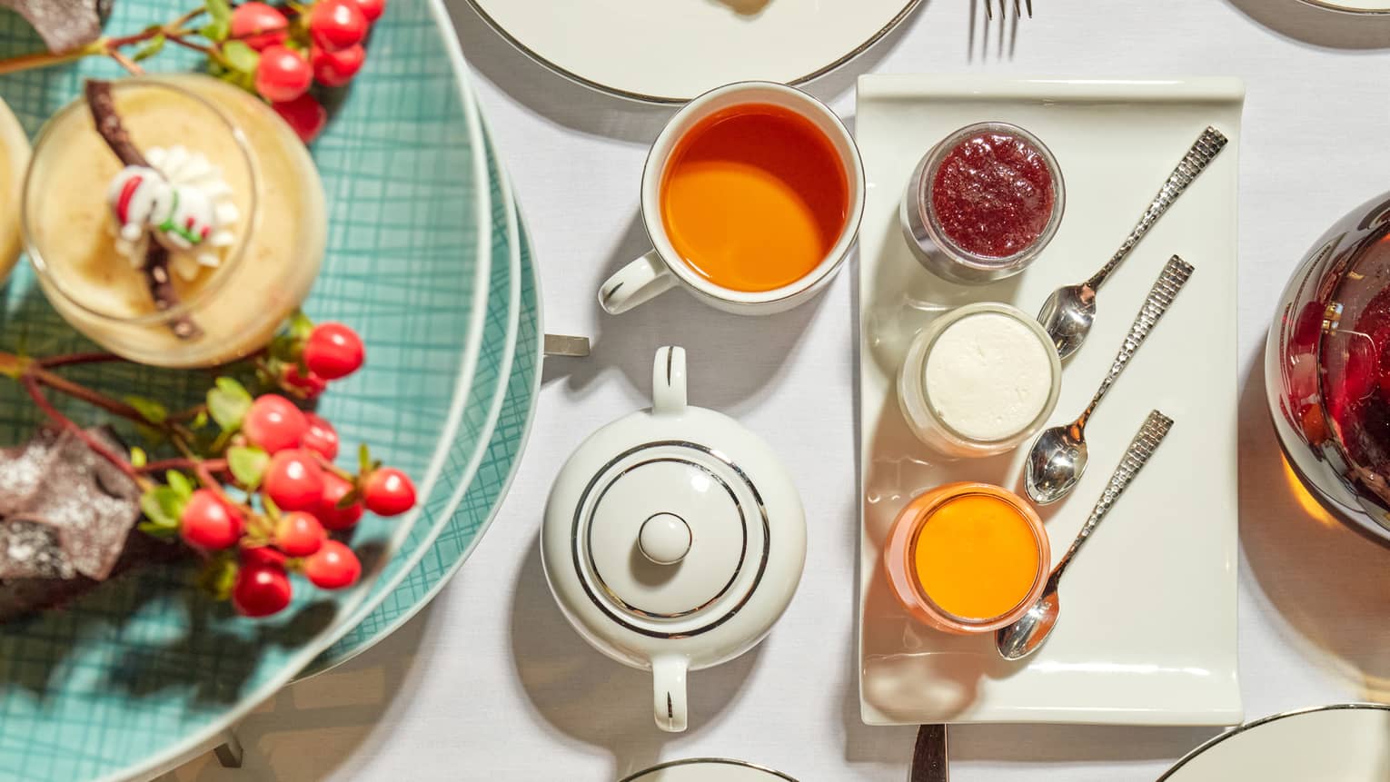 A guest drops a sugar cube into a cup on a table set with fine pastries, pots of jam and cream, cutlery and a tea service.