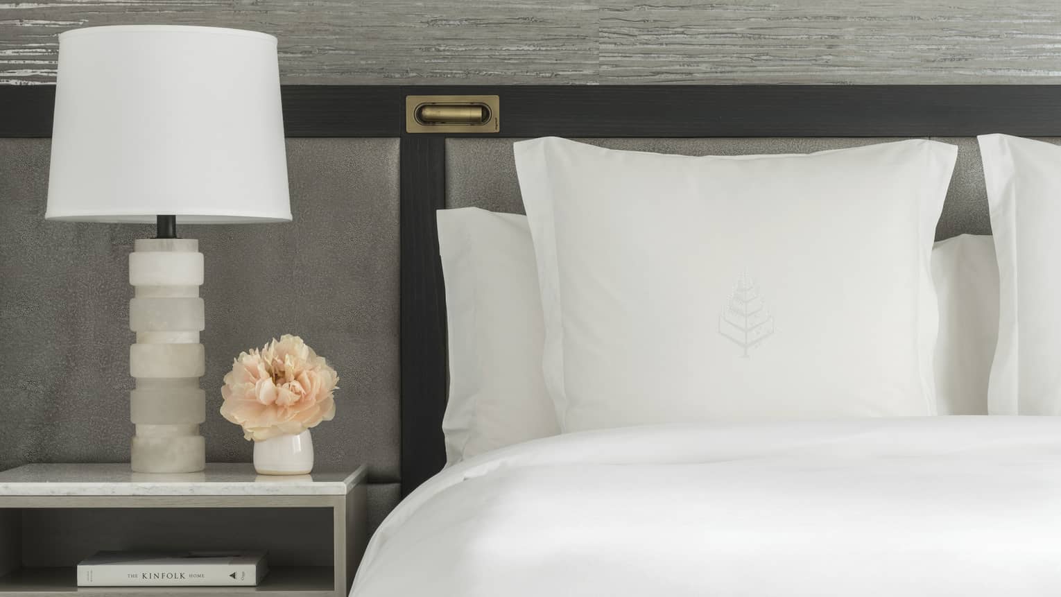 Luxury hotel room king bed with white sheets and Four Seasons tree logo embroidered on sham, next to a nightstand with white lamp and small vase of peach-coloured flowers