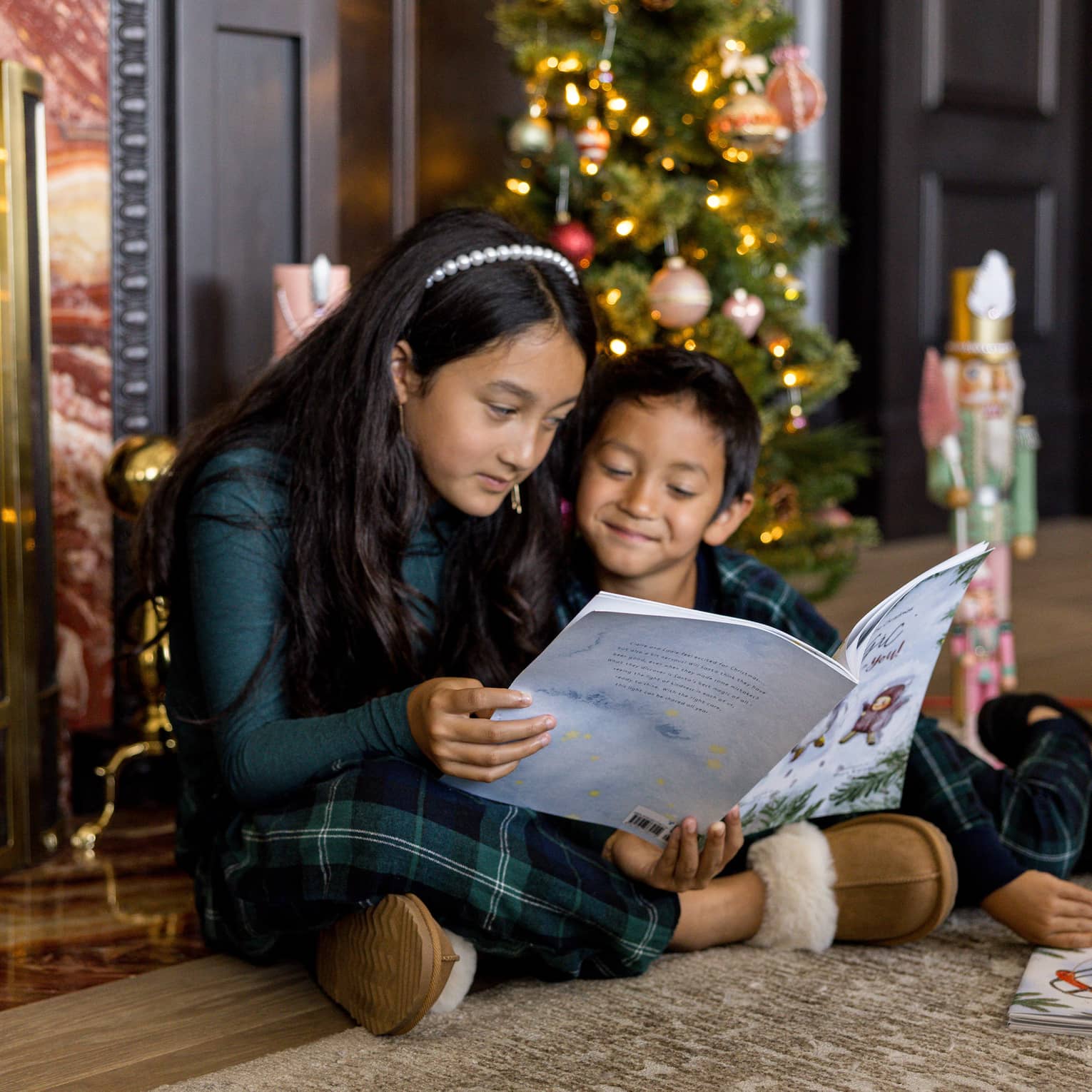 Two children wearing blue-and-green plaid pajamas sit in front of a fireplace and Christmas tree reading a book