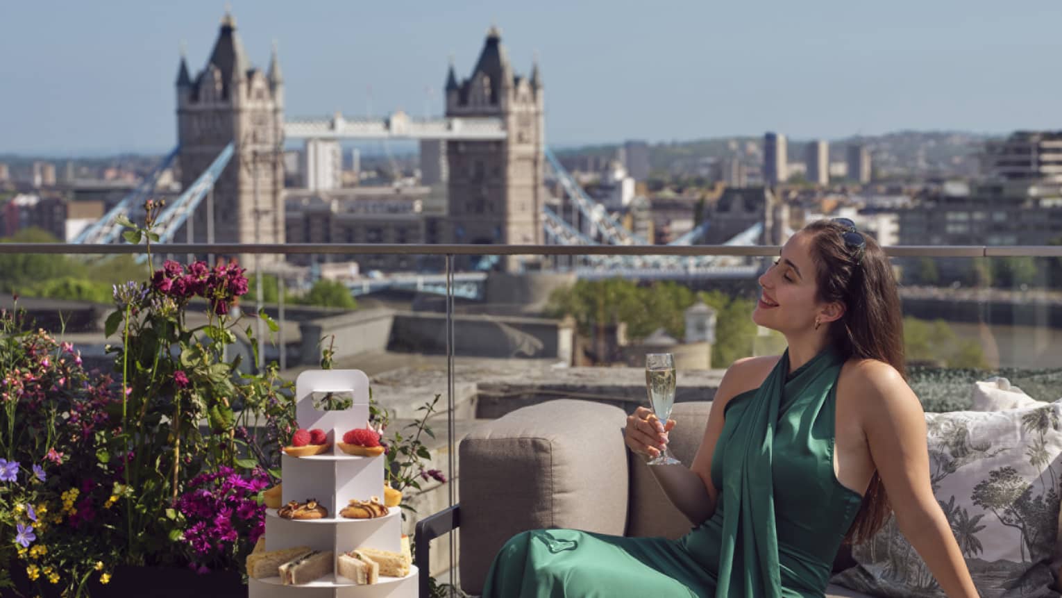 Woman in green dress holding champagne beside tea stand with confections and Tower Bridge in backdrop
