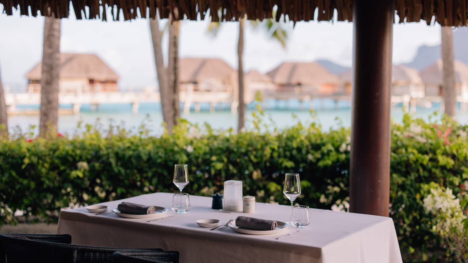An elegant table for two, separated from the ocean by a flowering hedge. Grass-roofed suites on stilts stand in the water.