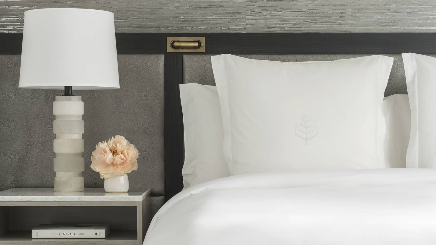 Luxury hotel room king bed with white sheets and Four Seasons tree logo embroidered on sham, next to a nightstand with white lamp and small vase of peach-coloured flowers