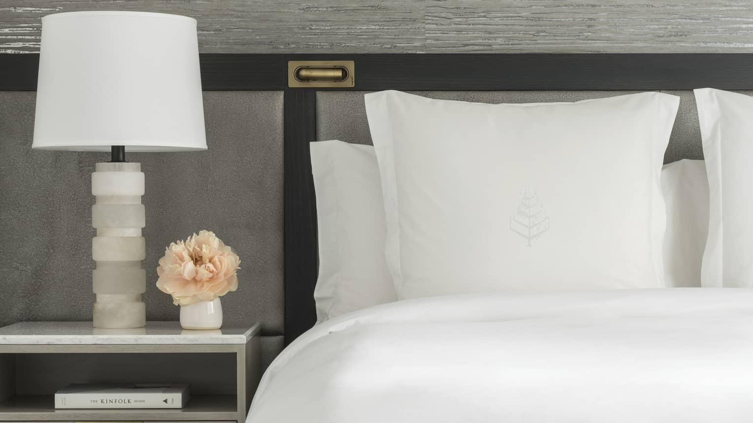 Luxury hotel room king bed with white sheets and Four Seasons tree logo embroidered on sham, next to a nightstand with white lamp and small vase of peach-coloured flowers