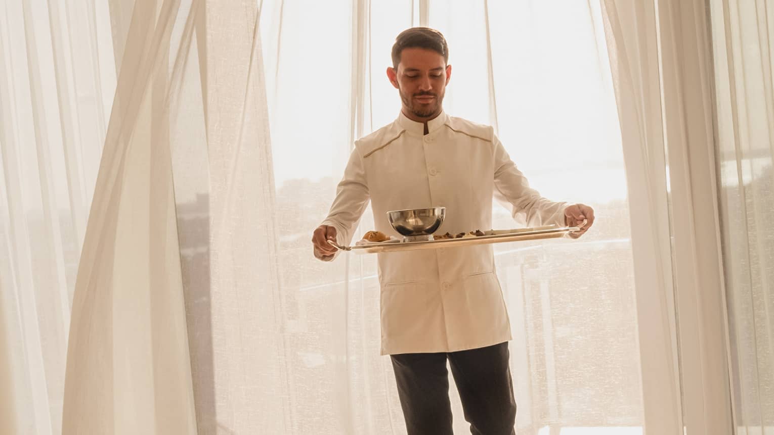 Hotel staff member carrying a tray with food for in-room dining, standing in front of sheer curtains with sunlight streaming through