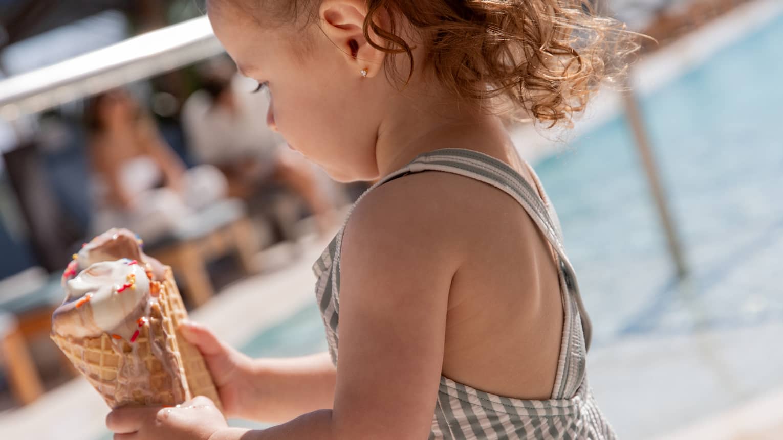 Toddler wearing a blue-and-white striped bathing suit carries an ice cream cone while walking by a hotel pool