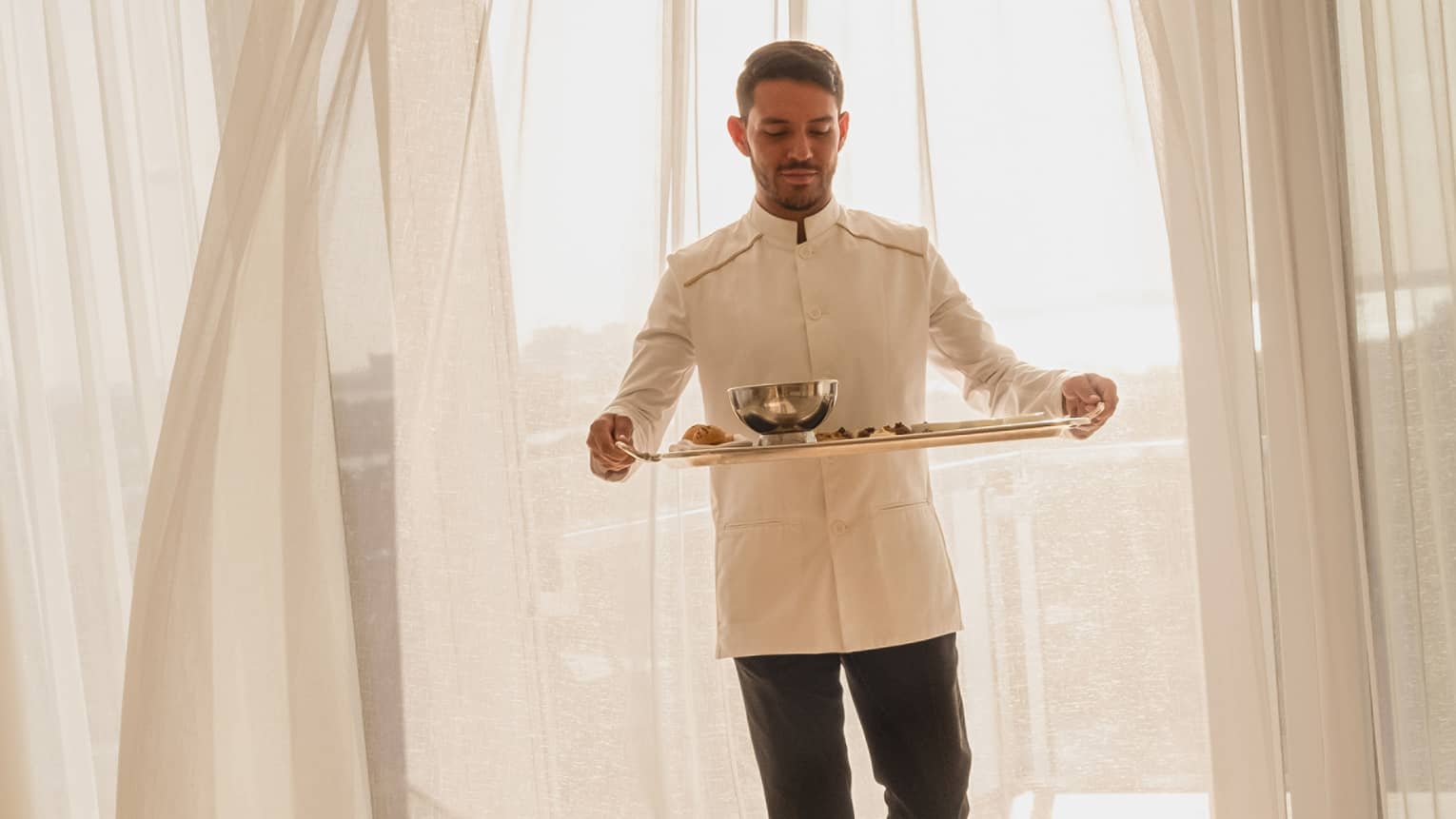 Hotel staff member carrying a tray with food for in-room dining, standing in front of sheer curtains with sunlight streaming through