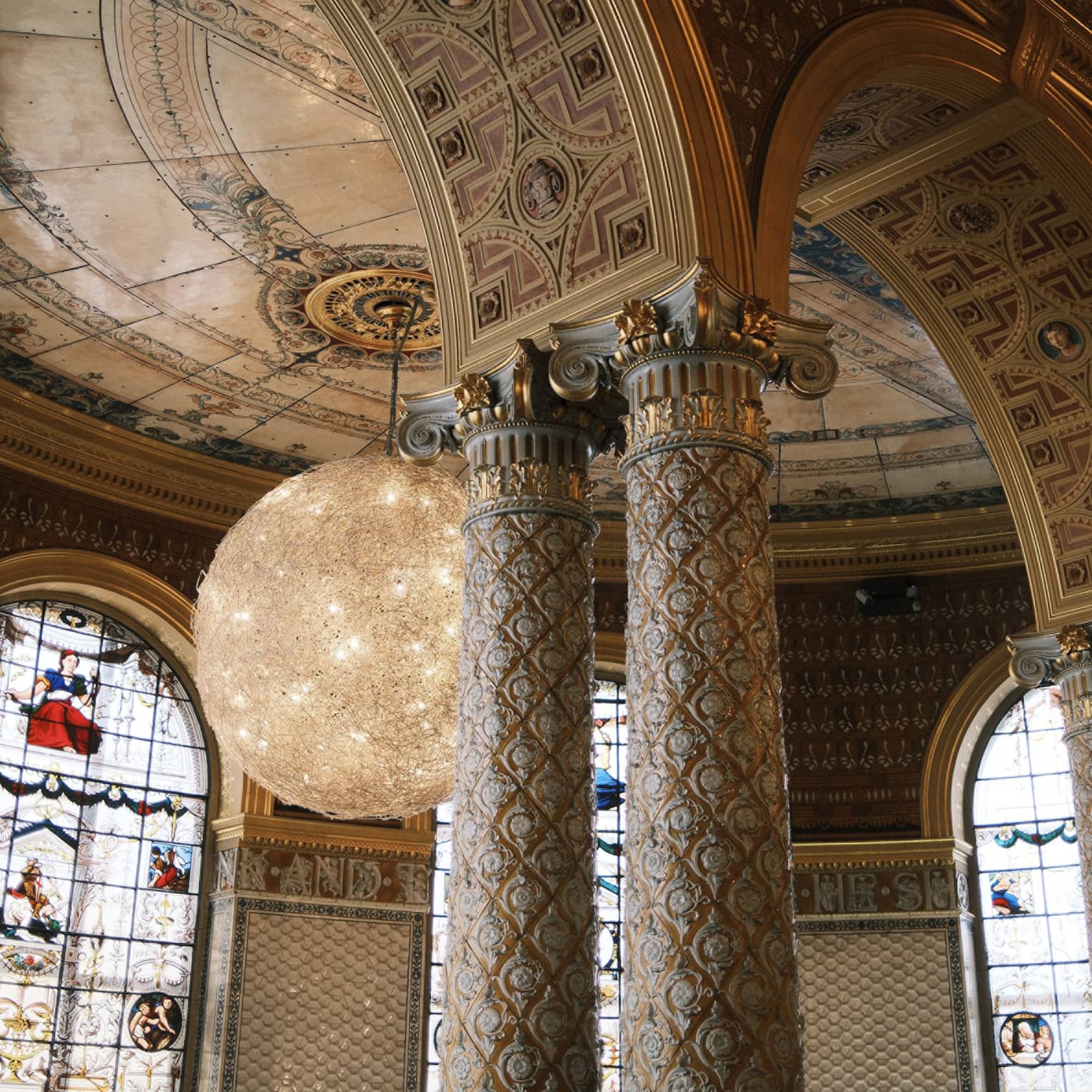 An interior space with a high arches and columns with decorative inlay and carving. Stained-glass windows, decorative ceiling and walls, and chandelier lighting are in the background.