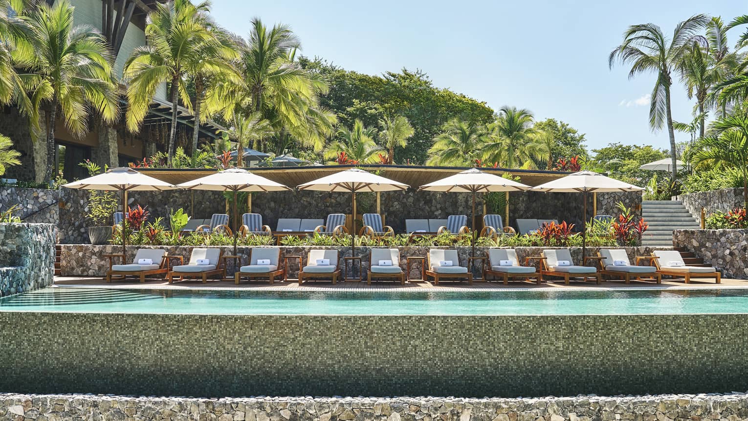 A line of lounge chairs and umbrellas at an outdoor pool surrounded by lush greenery