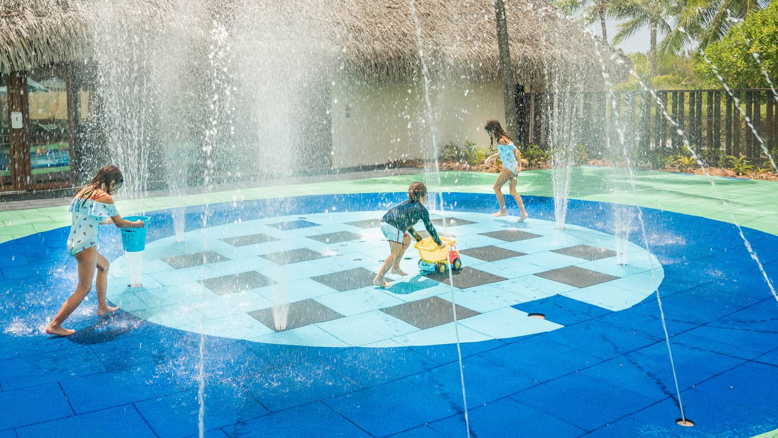 Group of children in swimsuits play on splash pad next to grass-roof building
