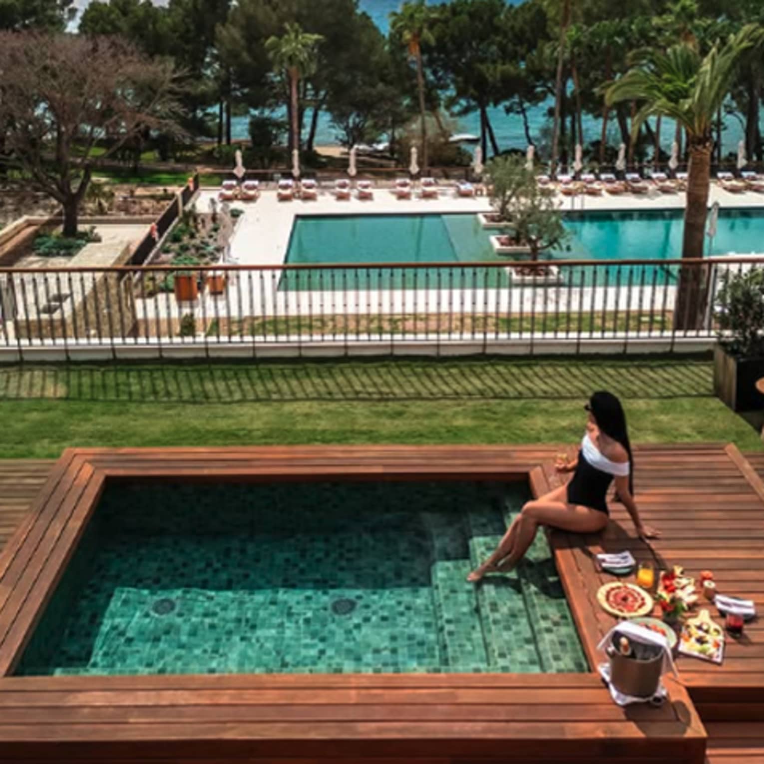 A woman lounges at a plunge pool with various food dishes, beverages and champagne, overlooking a large pool area and water view