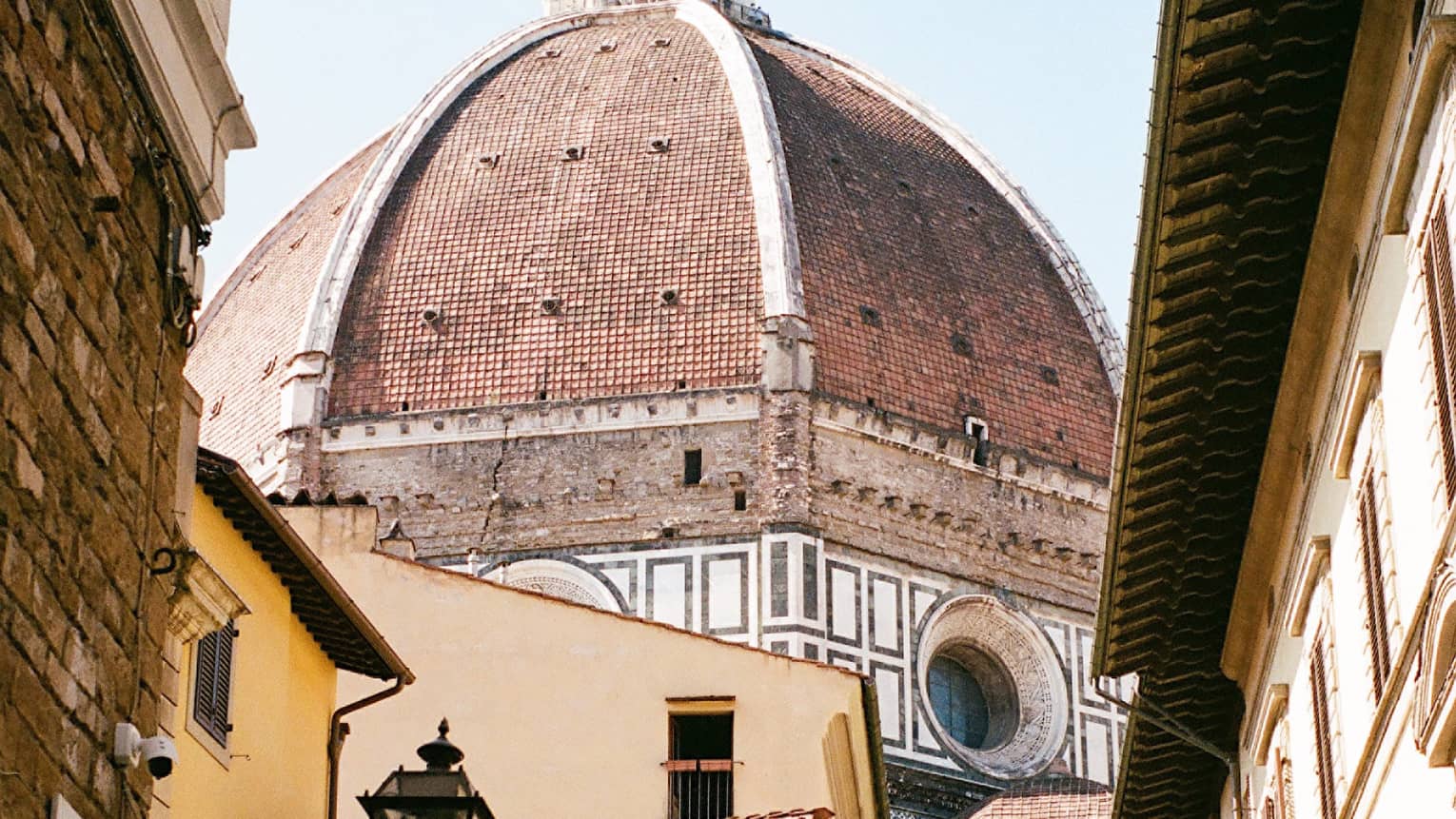 Looking up at the Florence Cathedral's dome from a narrow street below, a bright yellow shop building in the foreground.