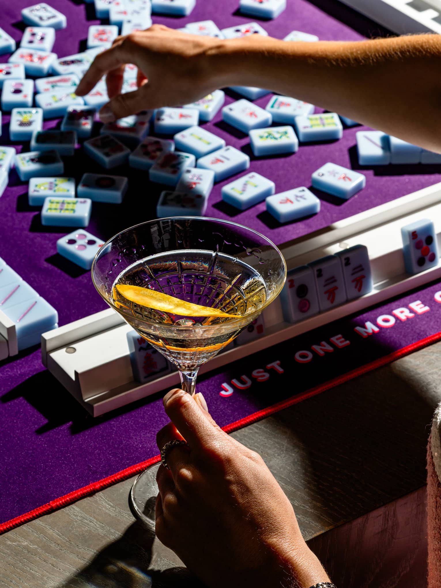 Close up of a person's hands, with one had reaching for a mahjong tile and the other holding a cocktail