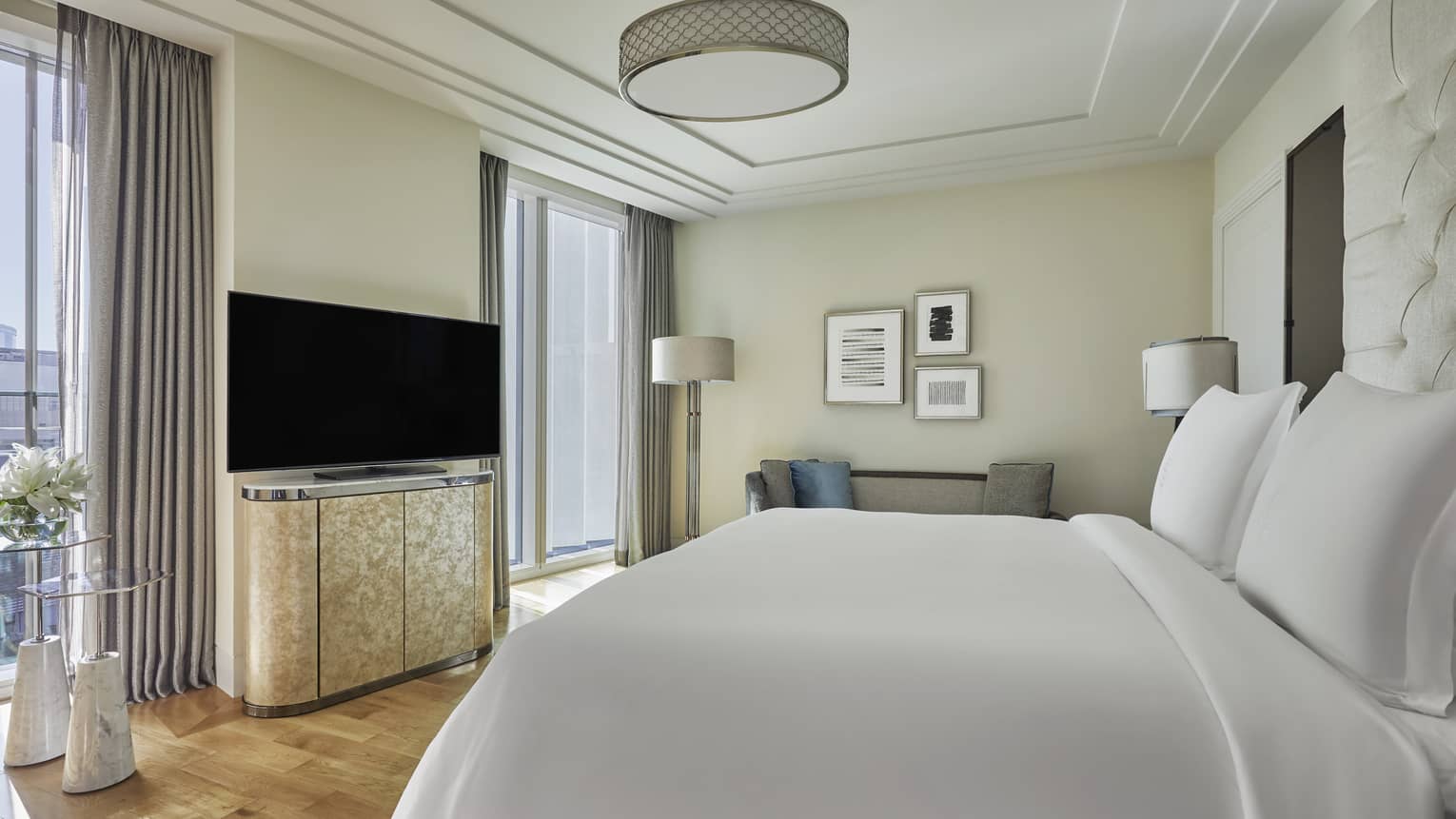 Bright hotel bedroom with a plush white bed, a flat-screen TV on a sleek cabinet, modern decor and large windows with gray curtains