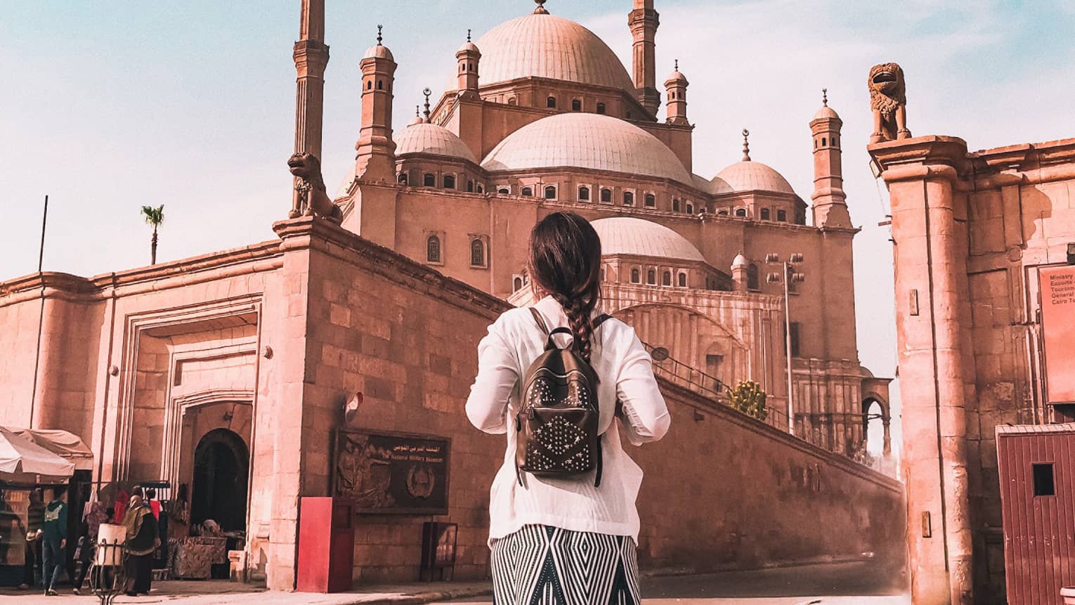 A tourist in a long skirt faces away, observing the Mosque of Muhammad Ali that towers over a wall surrounding it.