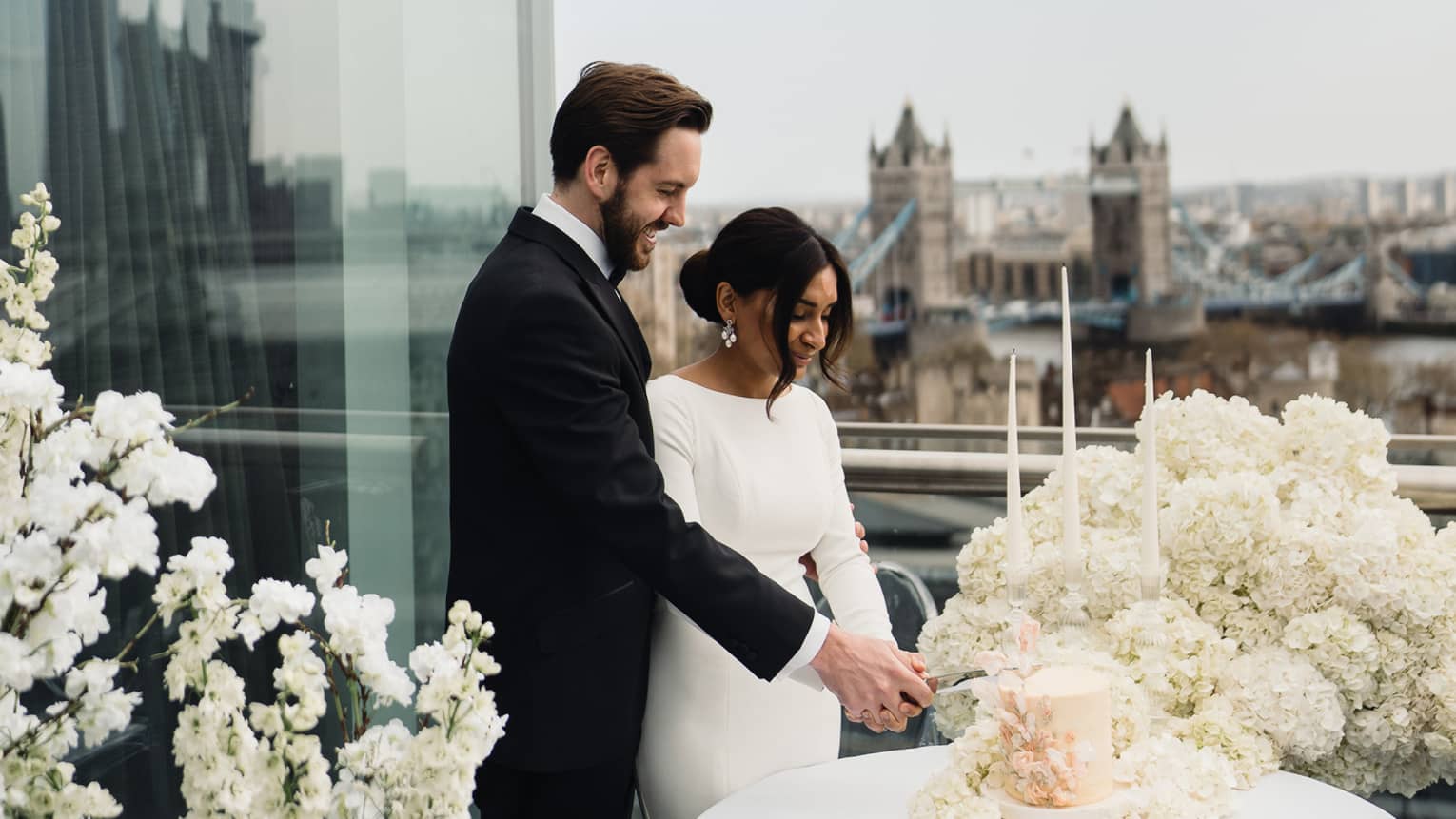 Wedding couple on a terrace cutting a pink wedding cake amid white flowers together