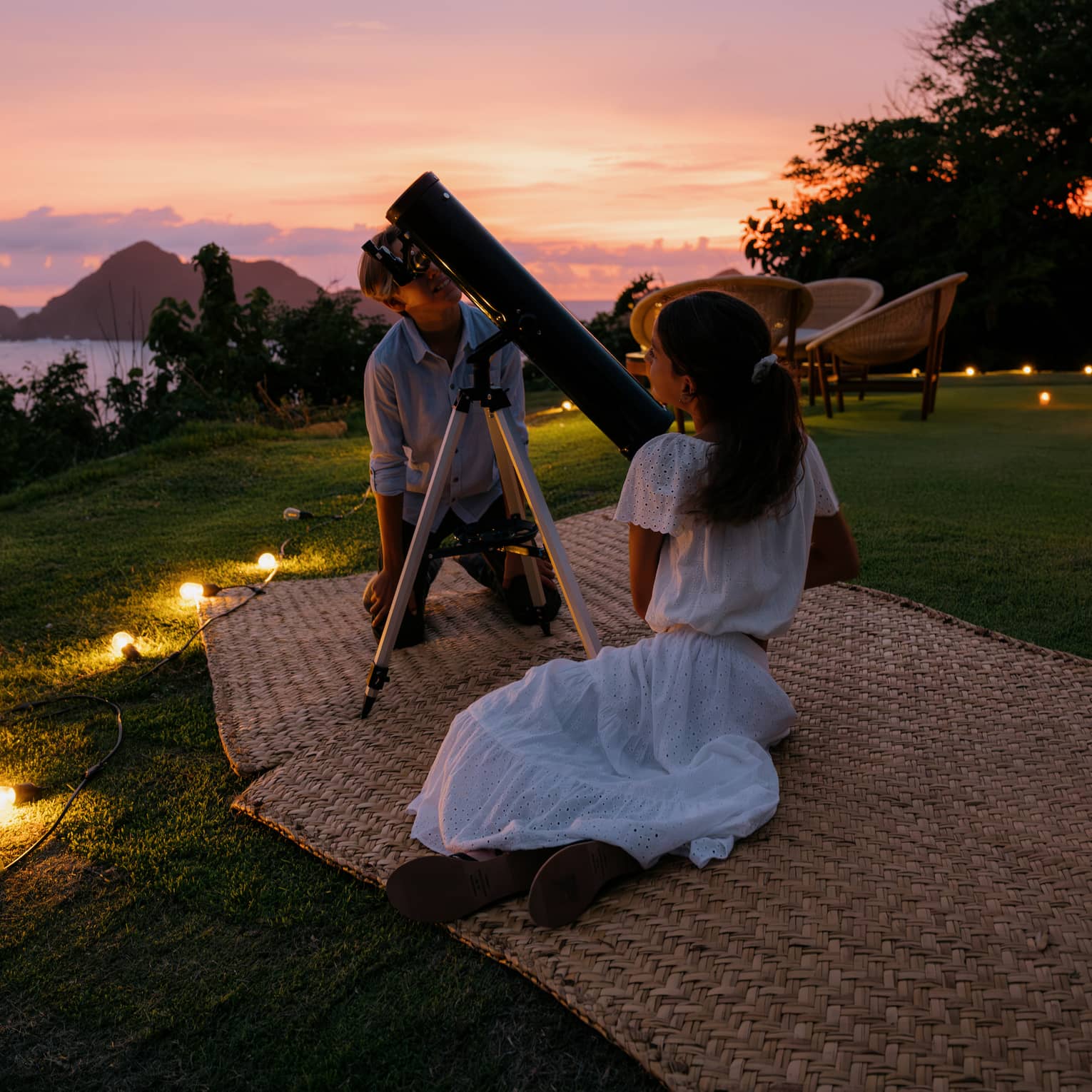 Two people on mats on the grass, with a large telescope. The ocean and a colorful sunset sky are in the background.