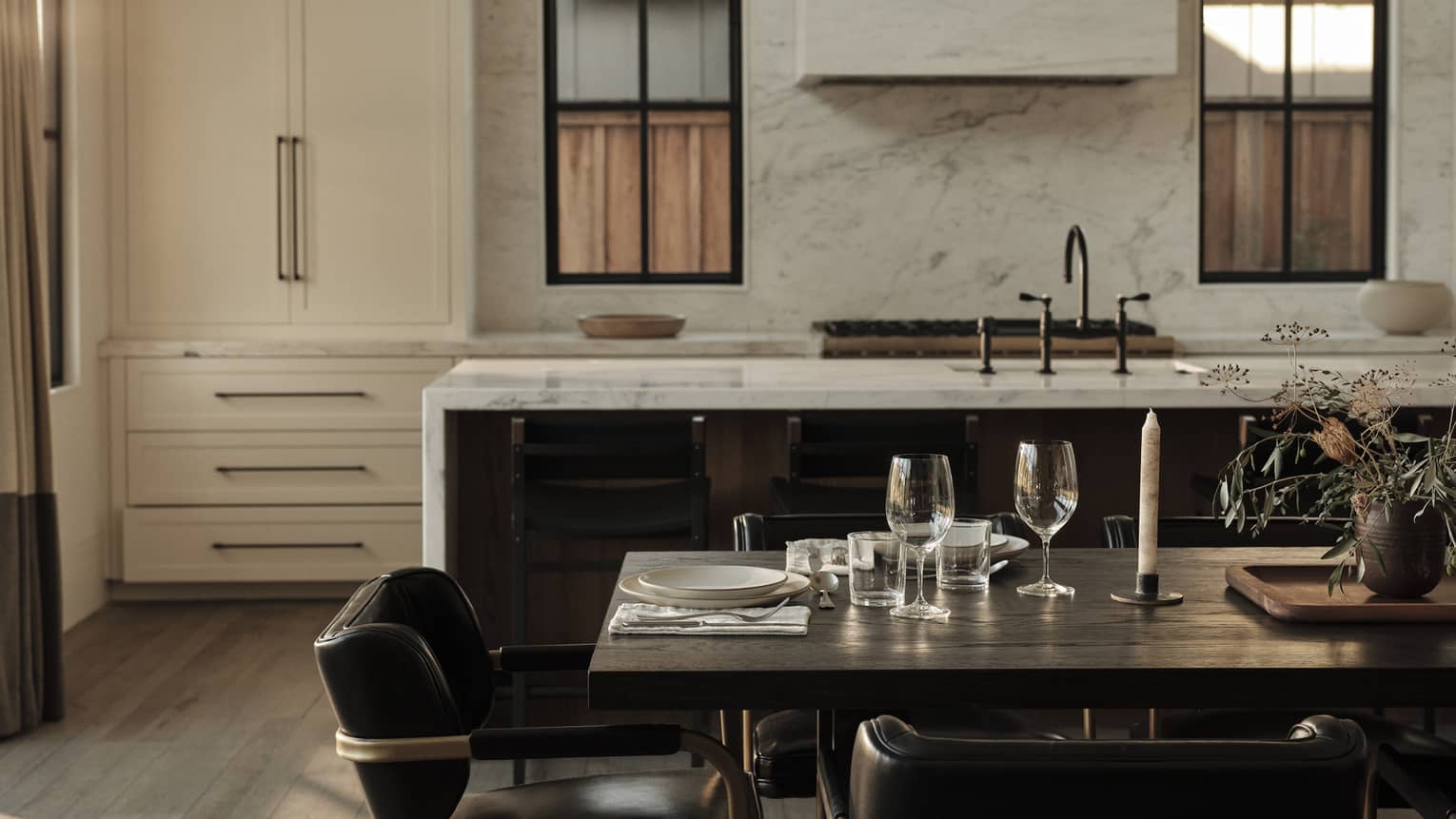 Luxury resort dining area set with glassware, plates and candles, overlooking a modern kitchen with marble island, dark chairs and large cabinets