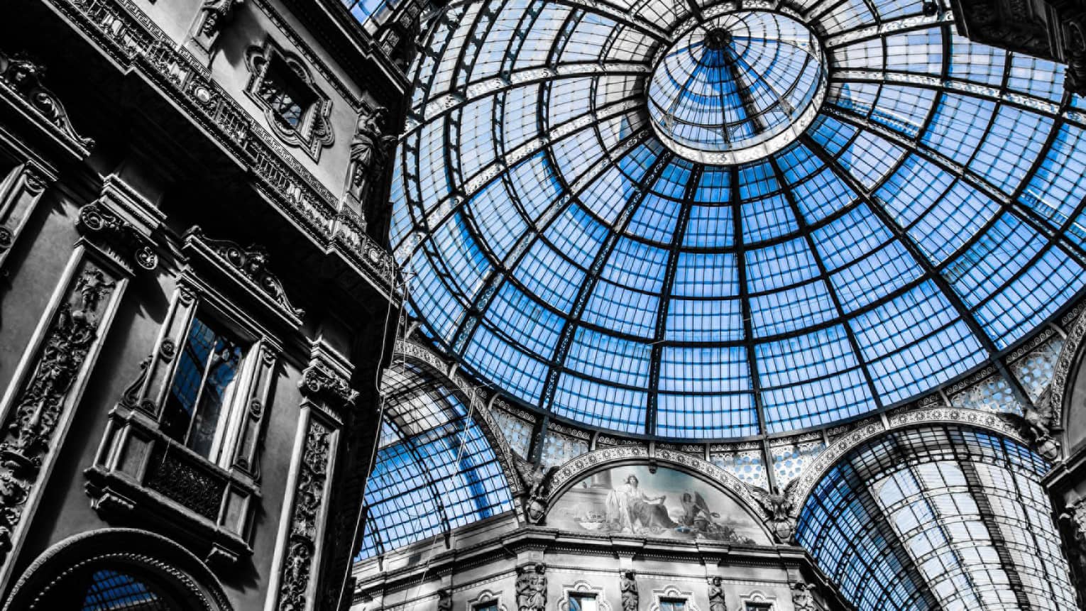 Interior view of the Galleria Vittorio Emanuele II in Milan, showcasing its grand glass dome ceiling and ornate architectural details in a mix of black and white with blue-tinted glass.