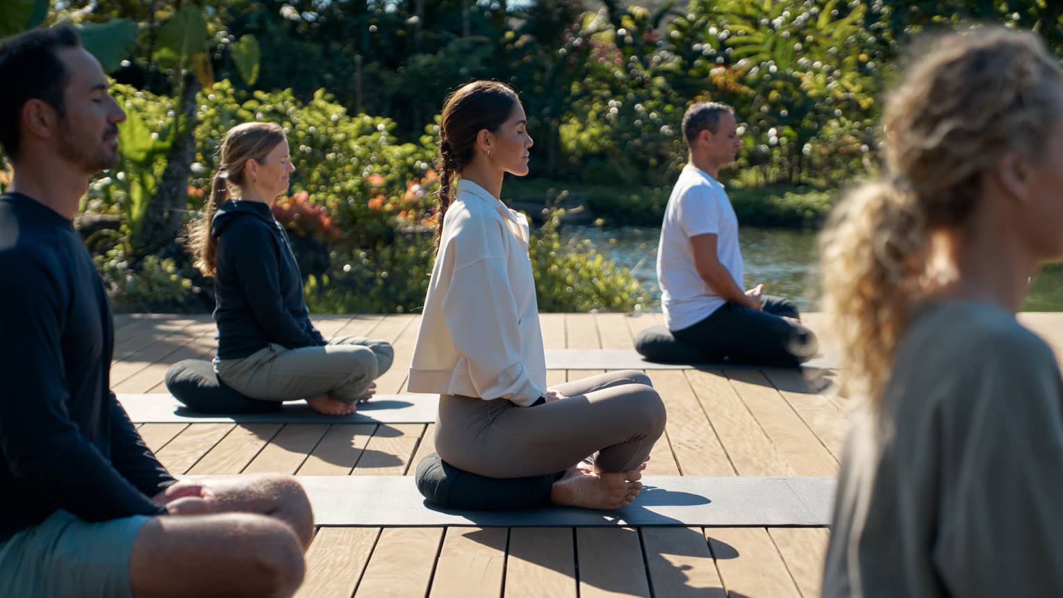 A group of people sit cross legged on wooden deck in garden setting, practicing yoga