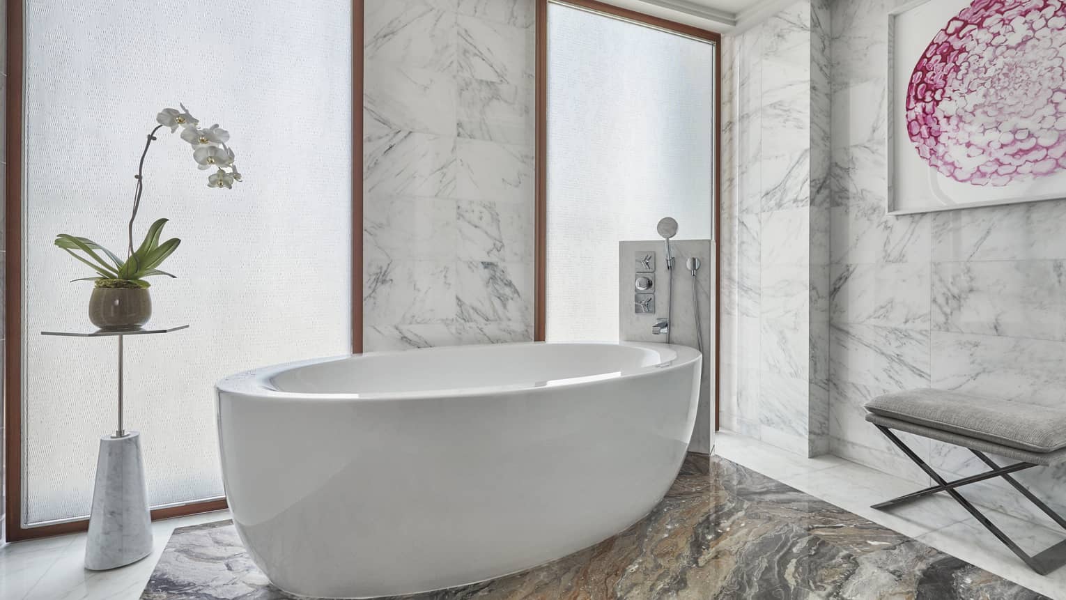 Luxurious marble bathroom featuring a freestanding oval bathtub, a modern orchid planter, textured glass windows and elegant decor accents