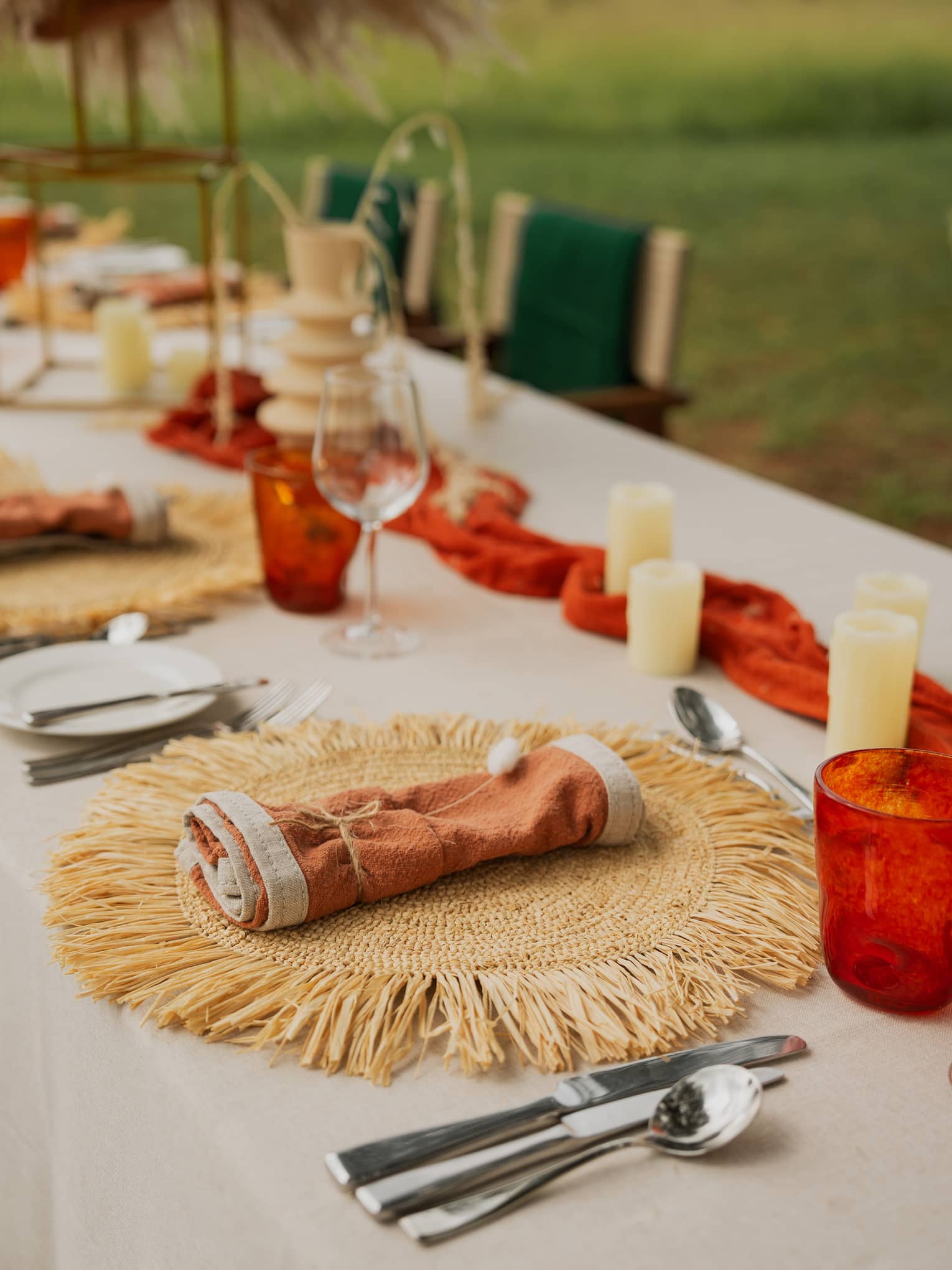Outdoor-inspired table setting with round rattan placemats, rust-coloured rolled napkins in centre, and orange and white votives lining the table centre