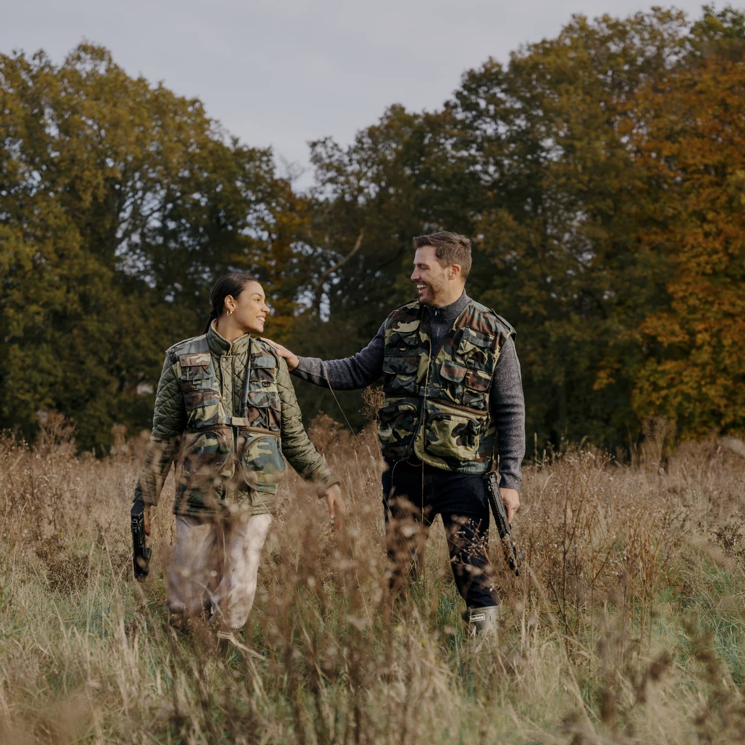 Two people wearing camouflage vests stand in a grassy field with trees in the background.