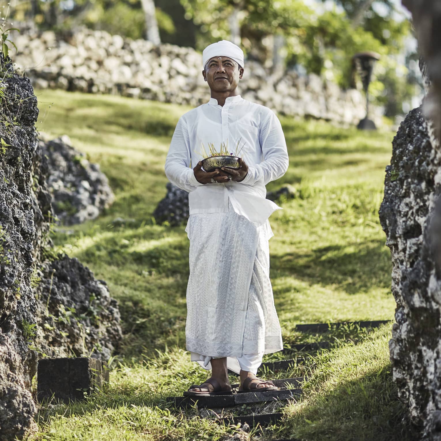 A priest in white attire stands on a stepping stone on a grassy hill, eyes gazing ahead, and holds a bowl of incense sticks.