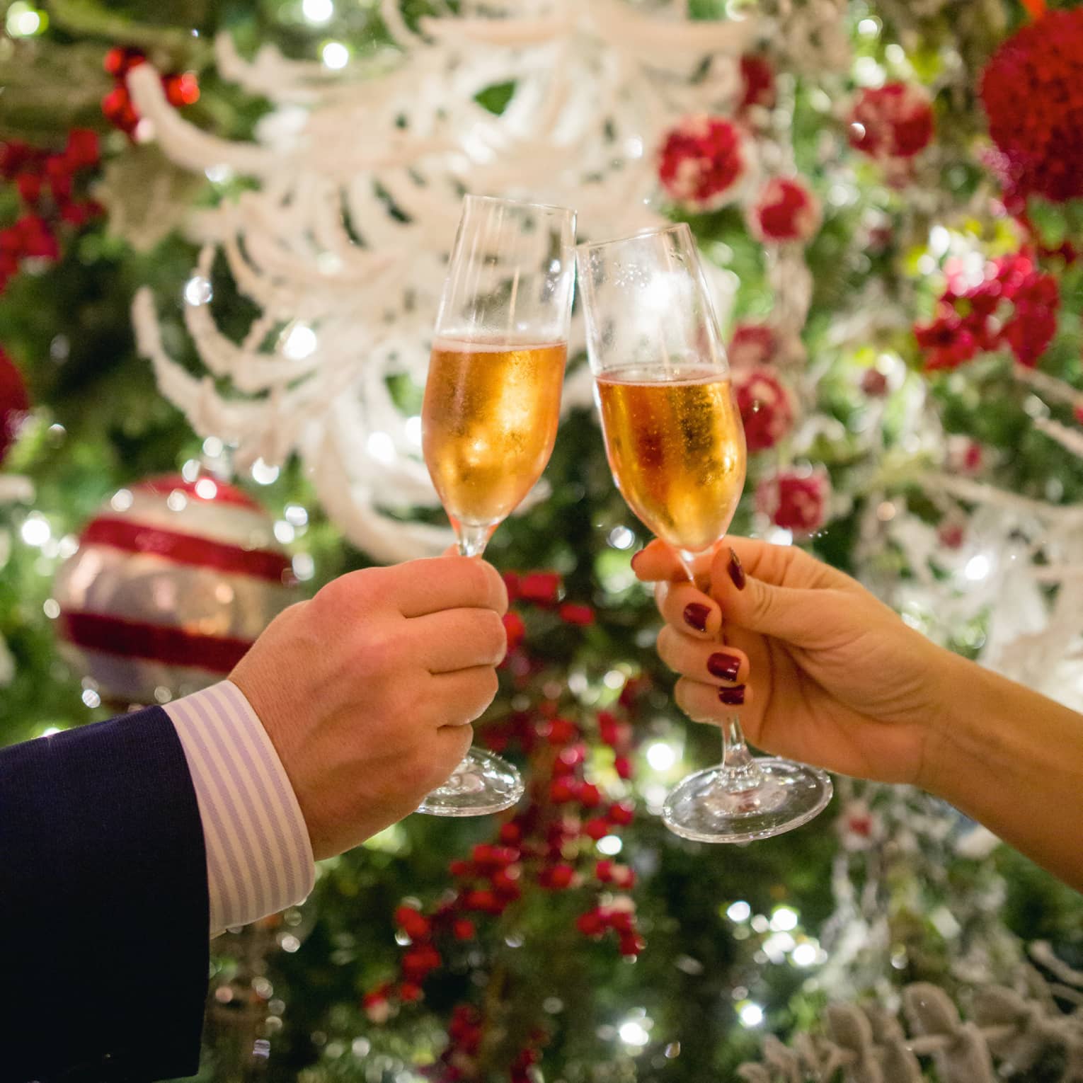 Two hands toasting Champagne glasses with holiday decor in backdrop
