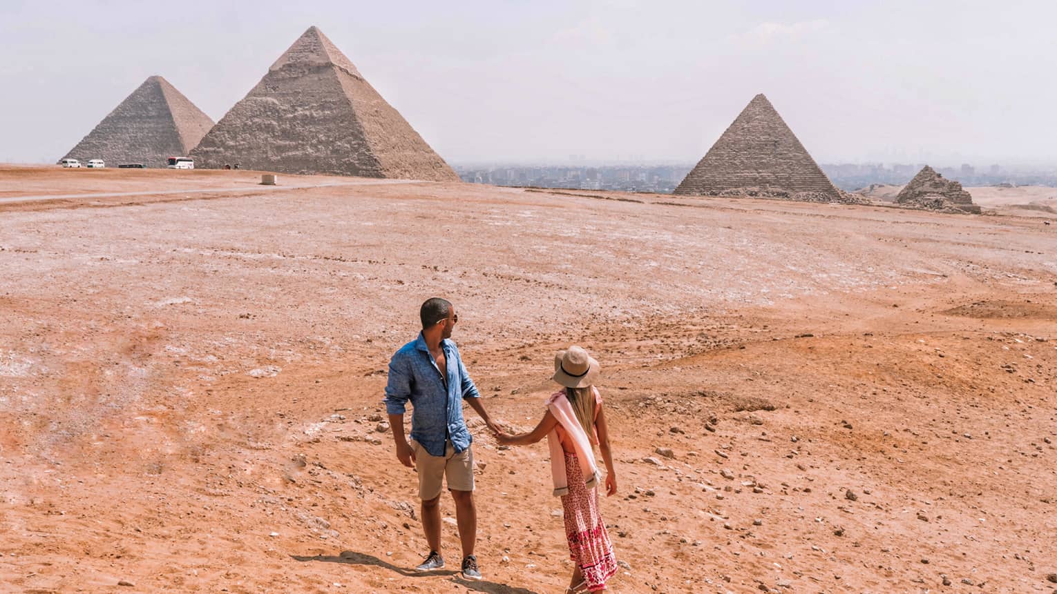 A couple holding hands turn back to look across the arid desert at the giant pyramids in the distance.