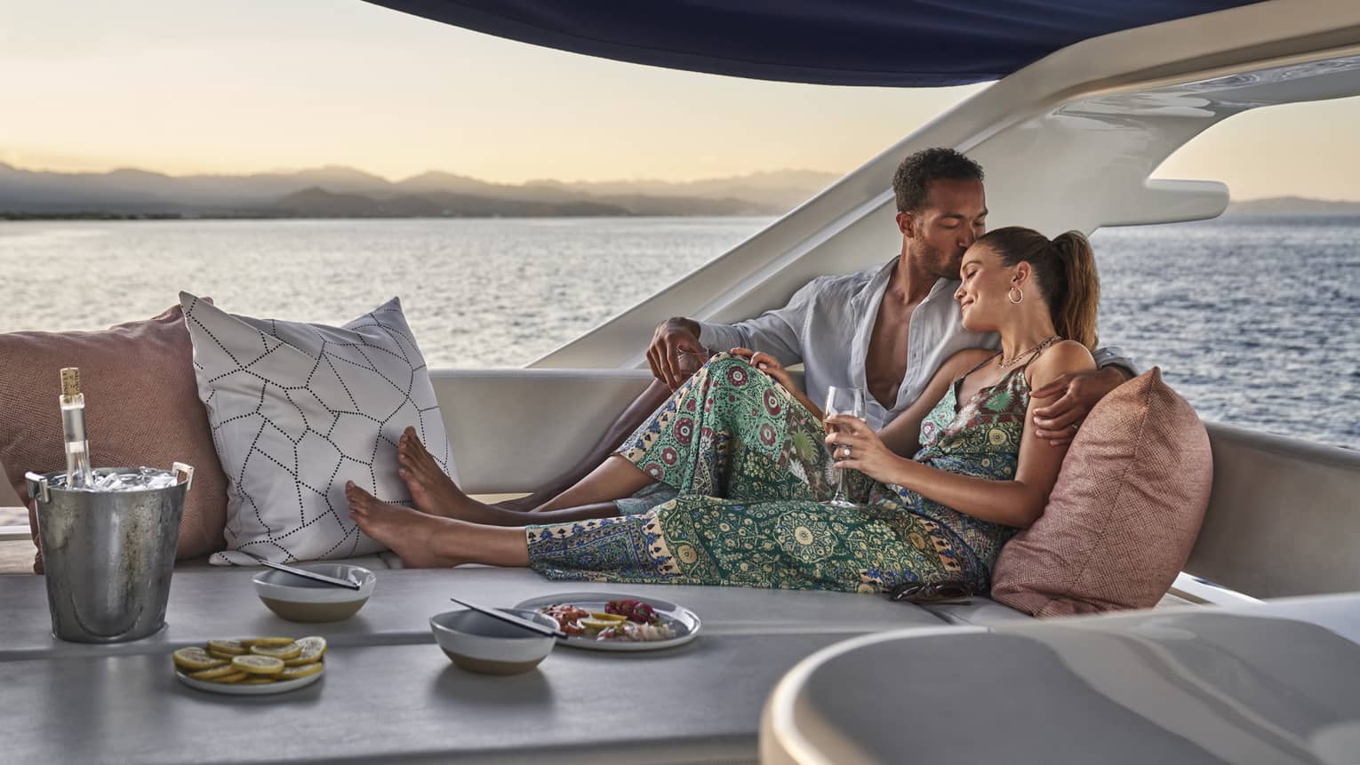 At sunset, a couple lounges in a luxury yacht set with a wine cooler, a plate of lemon slices and a dish of vibrant seafood.