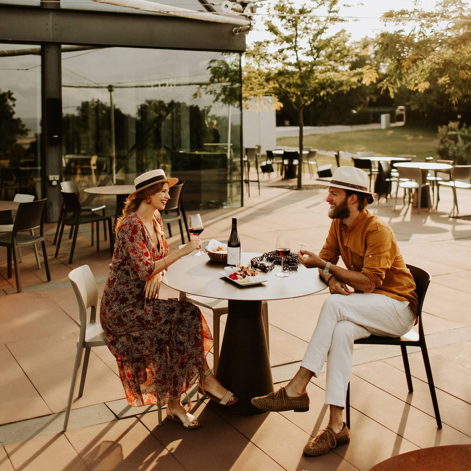 Couple wearing hats sit at outdoor table with plate of food, drinking red wine
