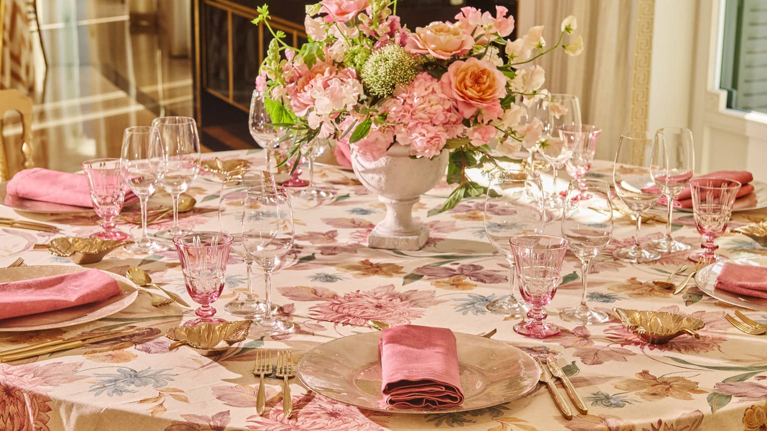 A table with floral tablecloth in pinkish hues with blue and gold accents is set with plates, golden leaf-shaped bowls, clear and pink-tinted stemware, pink napkins, utensils and a centerpiece of pink and white flowers in a white vase