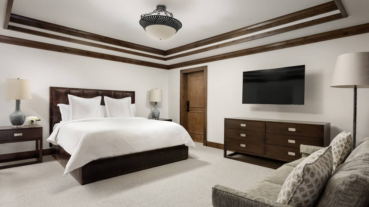Luxury bedroom with a king bed, dark leather headboard, two nightstands with lamps, wall-mounted TV, dresser and sofa