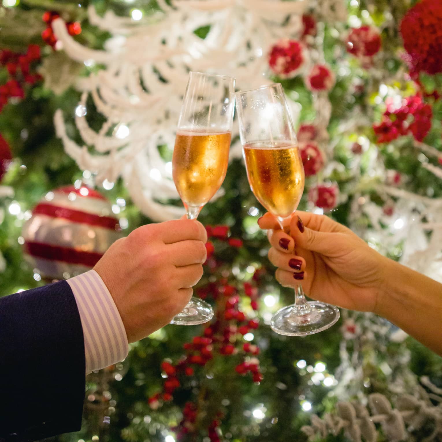 Two hands toasting Champagne glasses with holiday decor in backdrop