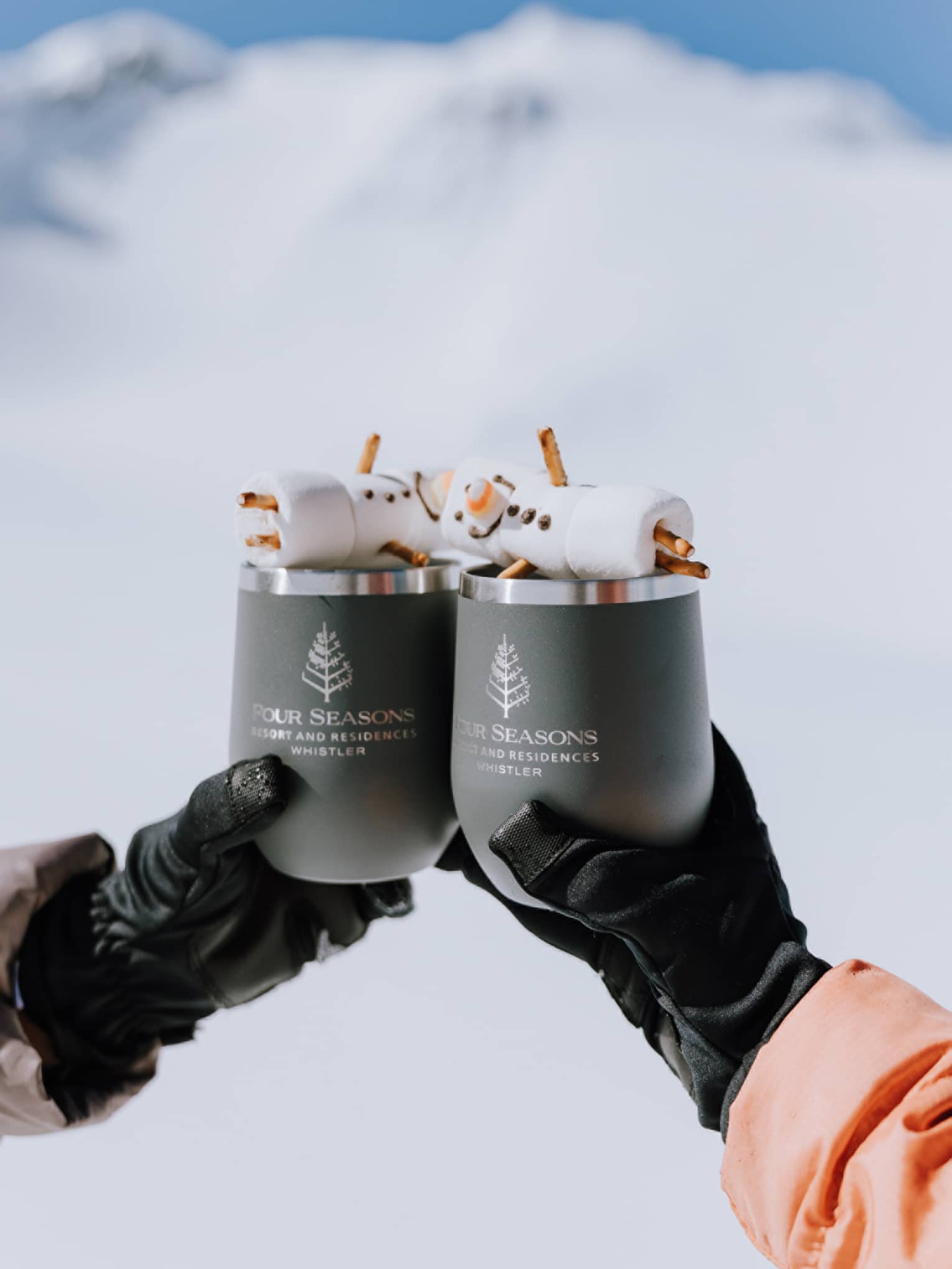 Two gloved hands each hold a mug with marshmallow snowmen on top