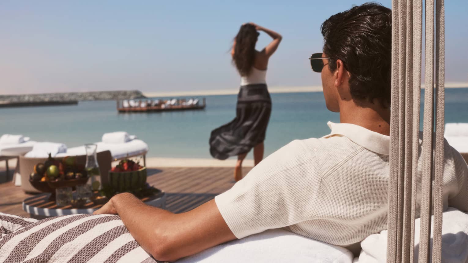 Man wearing white polo shirt lounges in a beachfront cabana while a woman wearing a white top and black maxi skirt walks toward the water