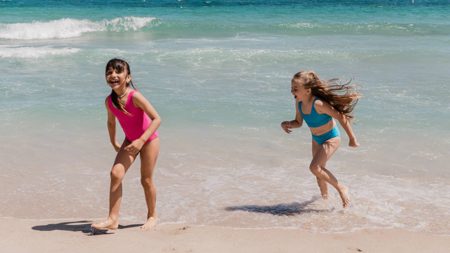 A young child wearing a bright pink bathing suit plays next to another child wearing a blue bathing suit at the edge of the water on the beach