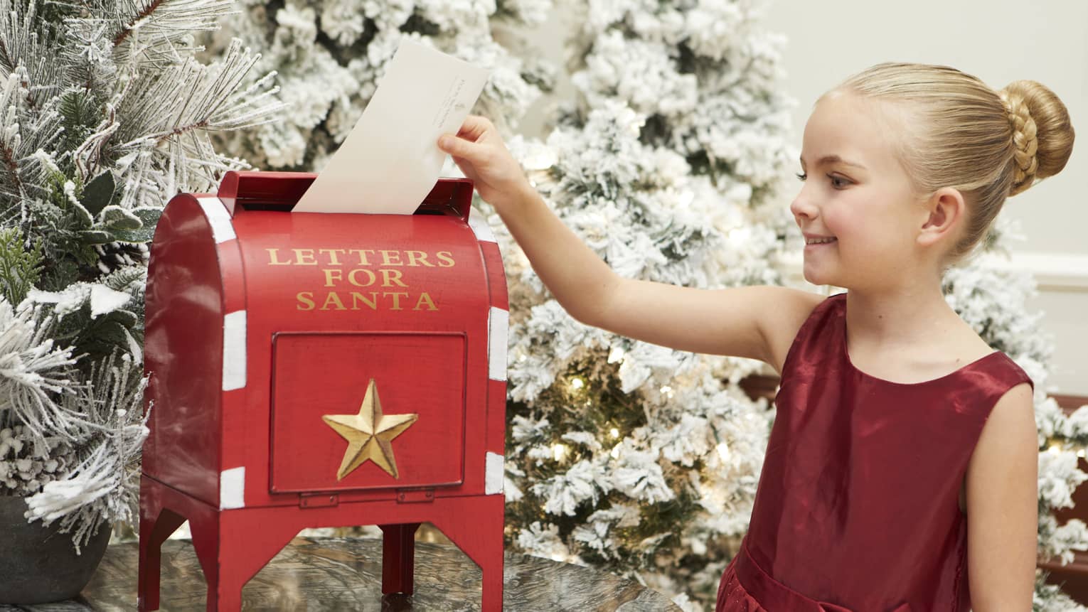 A young girl in a red dress putting a letter in a red box that says "For Santa."