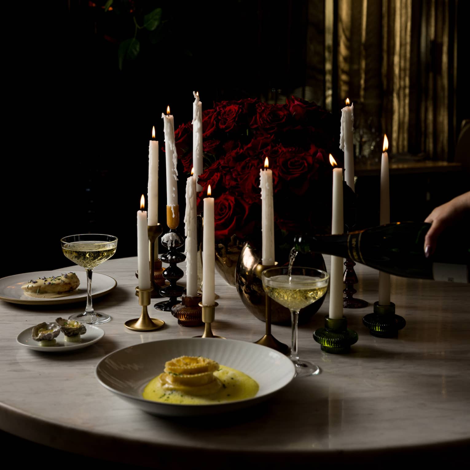 A round marble table topped with various pillar candles, a red rose centrepiece and food and drink for two is set in a dimly lit room