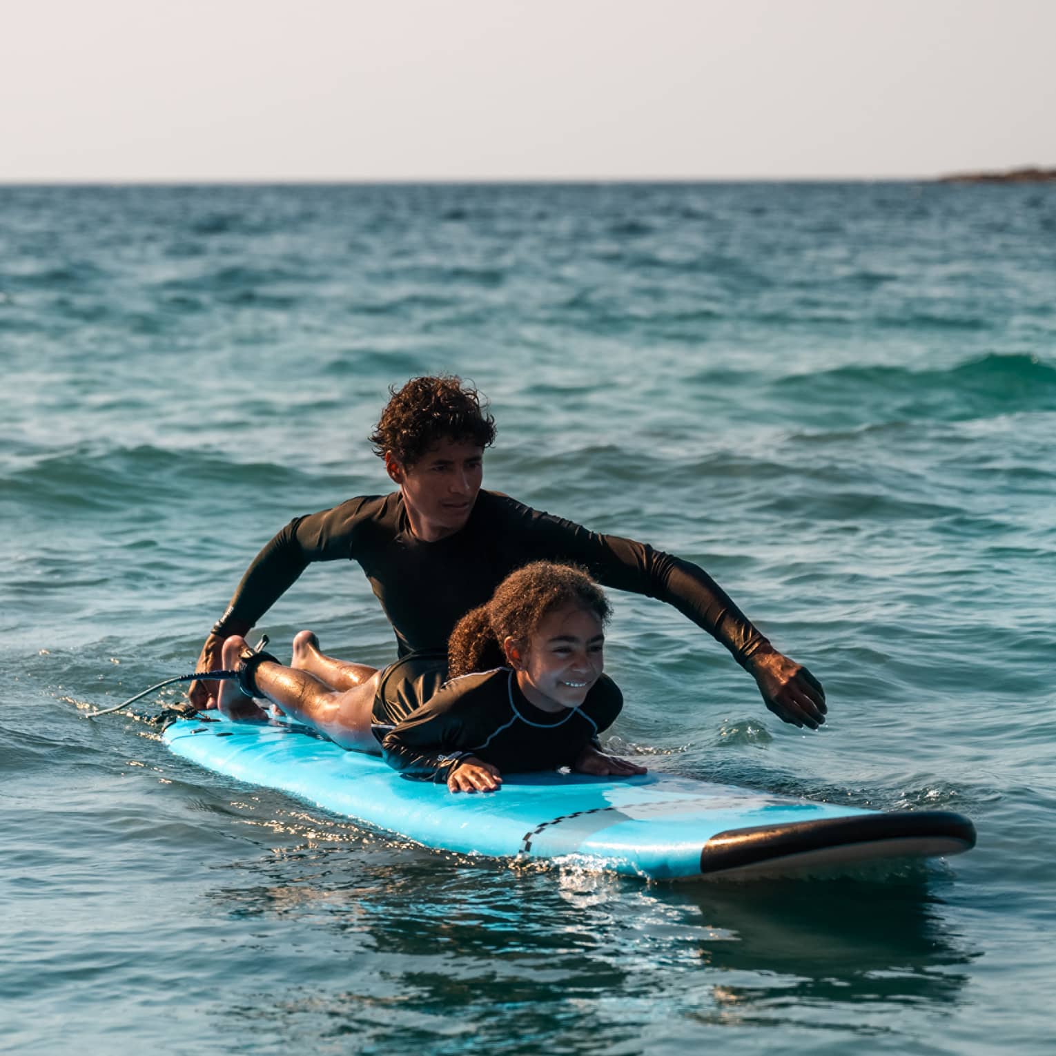 Male suf instructor wearing a black long-sleeve rashguard helps a young surfer paddle in the ocean