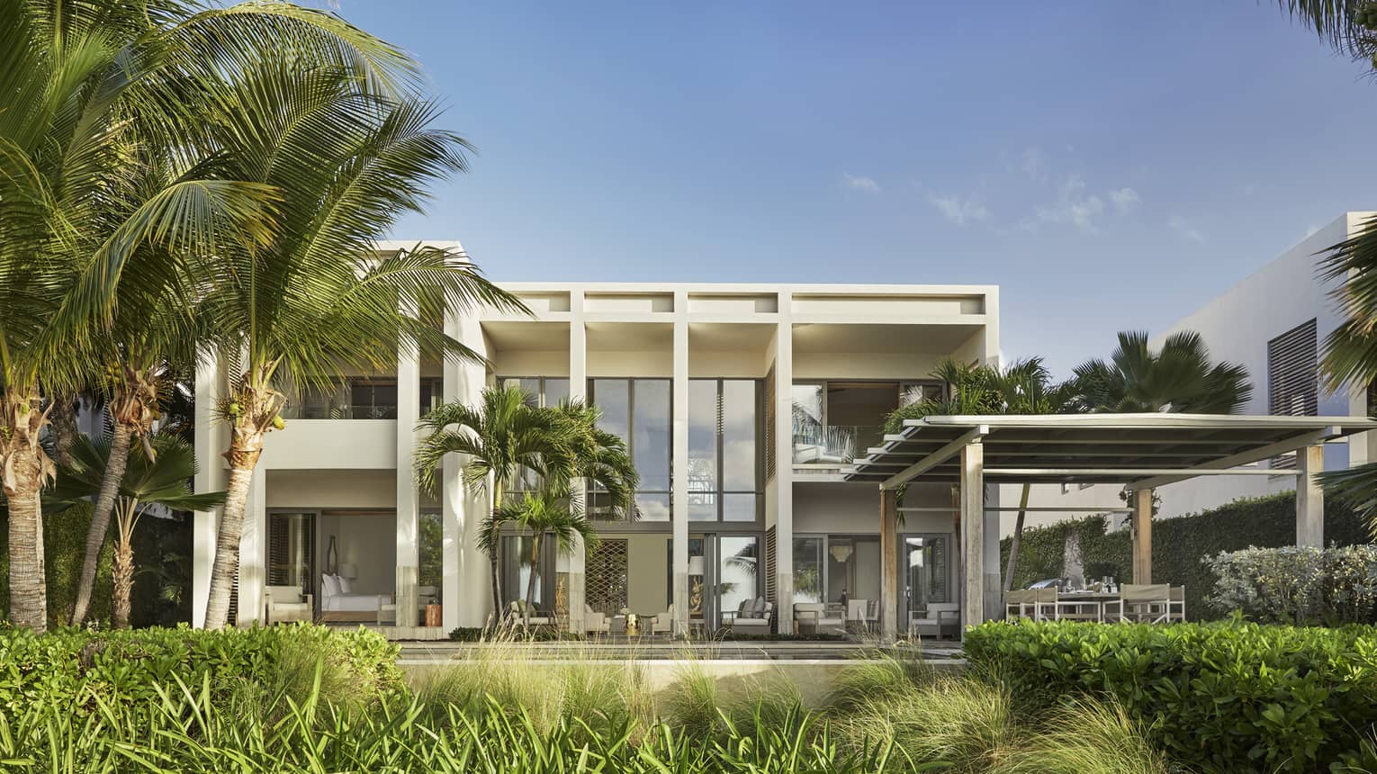 Modern beachfront villa with large glass windows, palm trees and a shaded outdoor seating area.